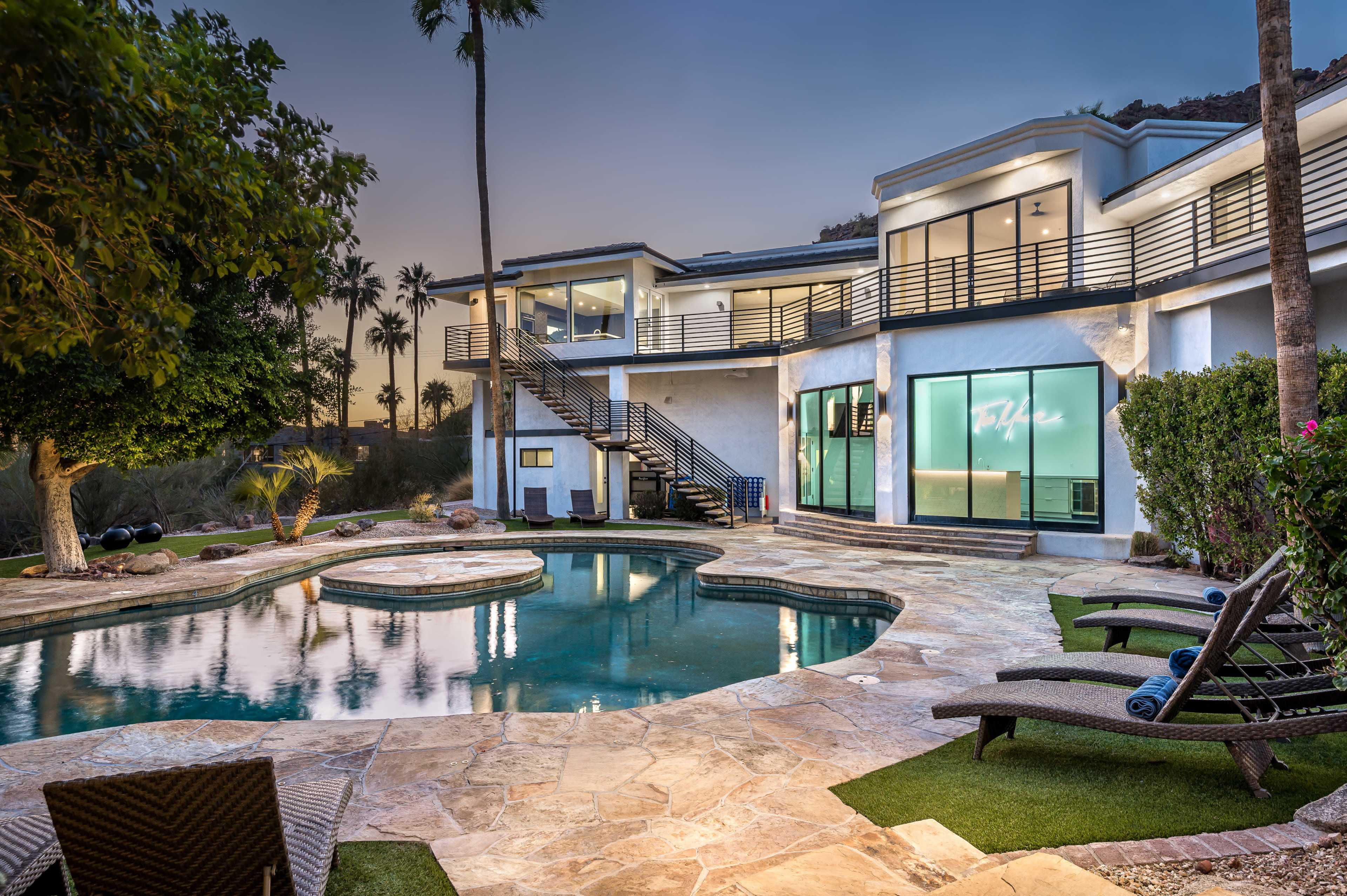 A modern house with large glass windows overlooks a swimming pool surrounded by stone patio and lounge chairs, set against a backdrop of palm trees and a twilight sky.
