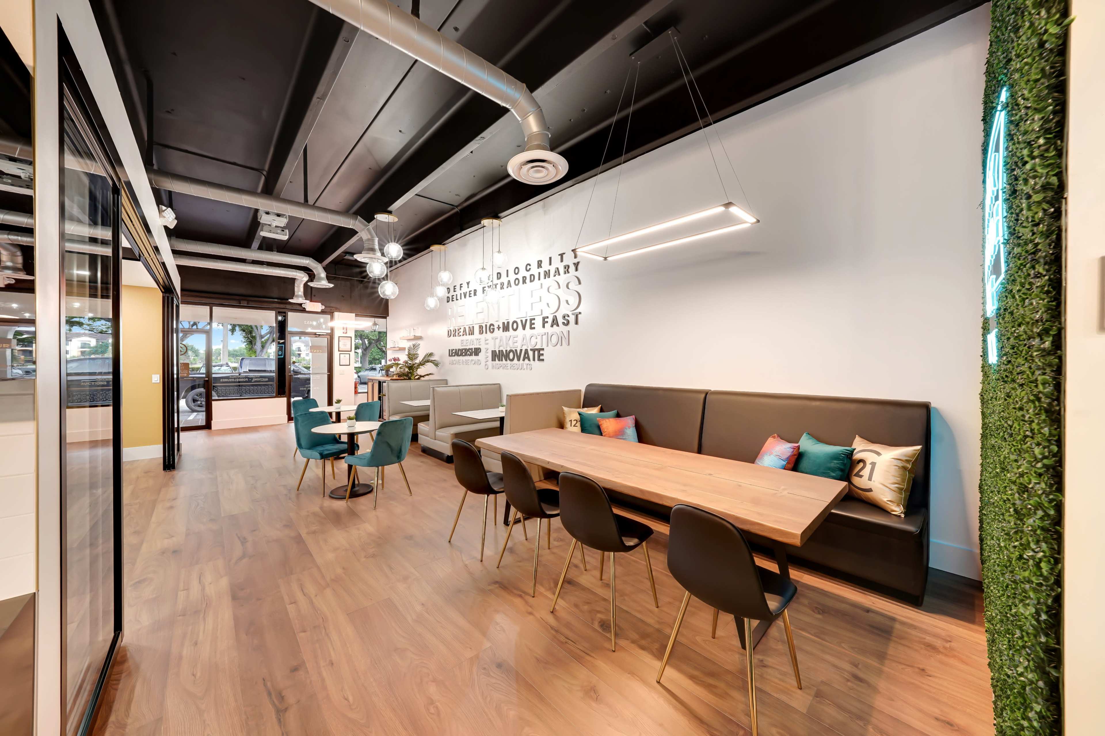 A modern office space featuring a dining area with a long wooden table and black chairs, flanked by a lounge area with colorful armchairs and a green wall element.