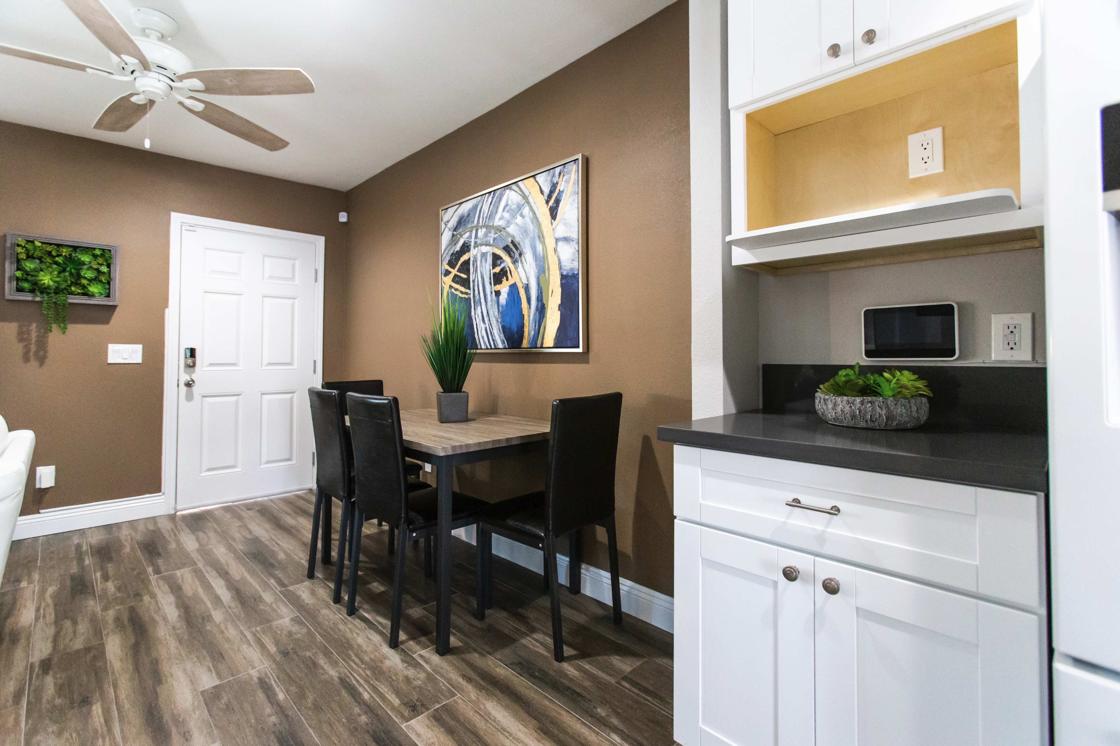 The image shows a dining area with a wooden table and four black chairs, adjacent to a kitchenette with white cabinetry and a microwave.