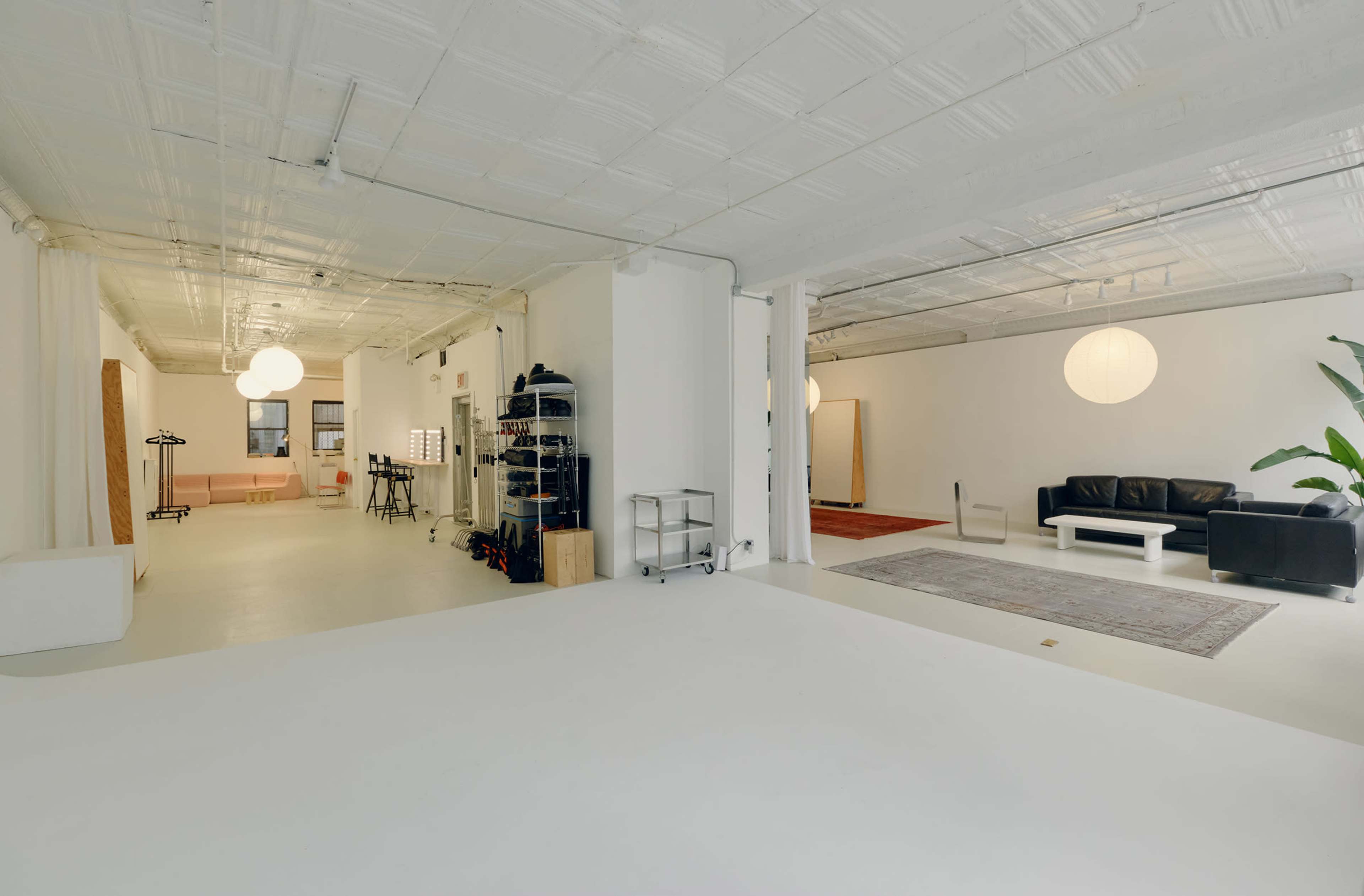 The image shows a spacious, well-lit studio with white walls, separate areas for lounging and work, and minimal furniture.