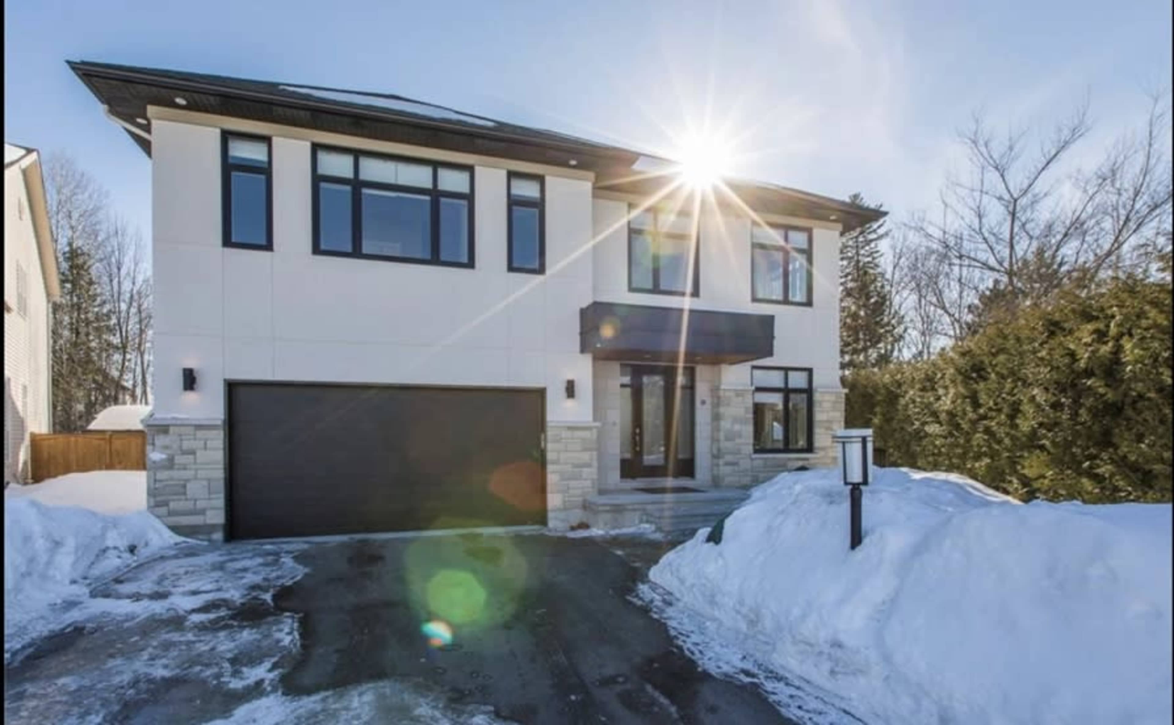 A modern two-story house with large windows and a driveway partially covered in snow is illuminated by sunlight.