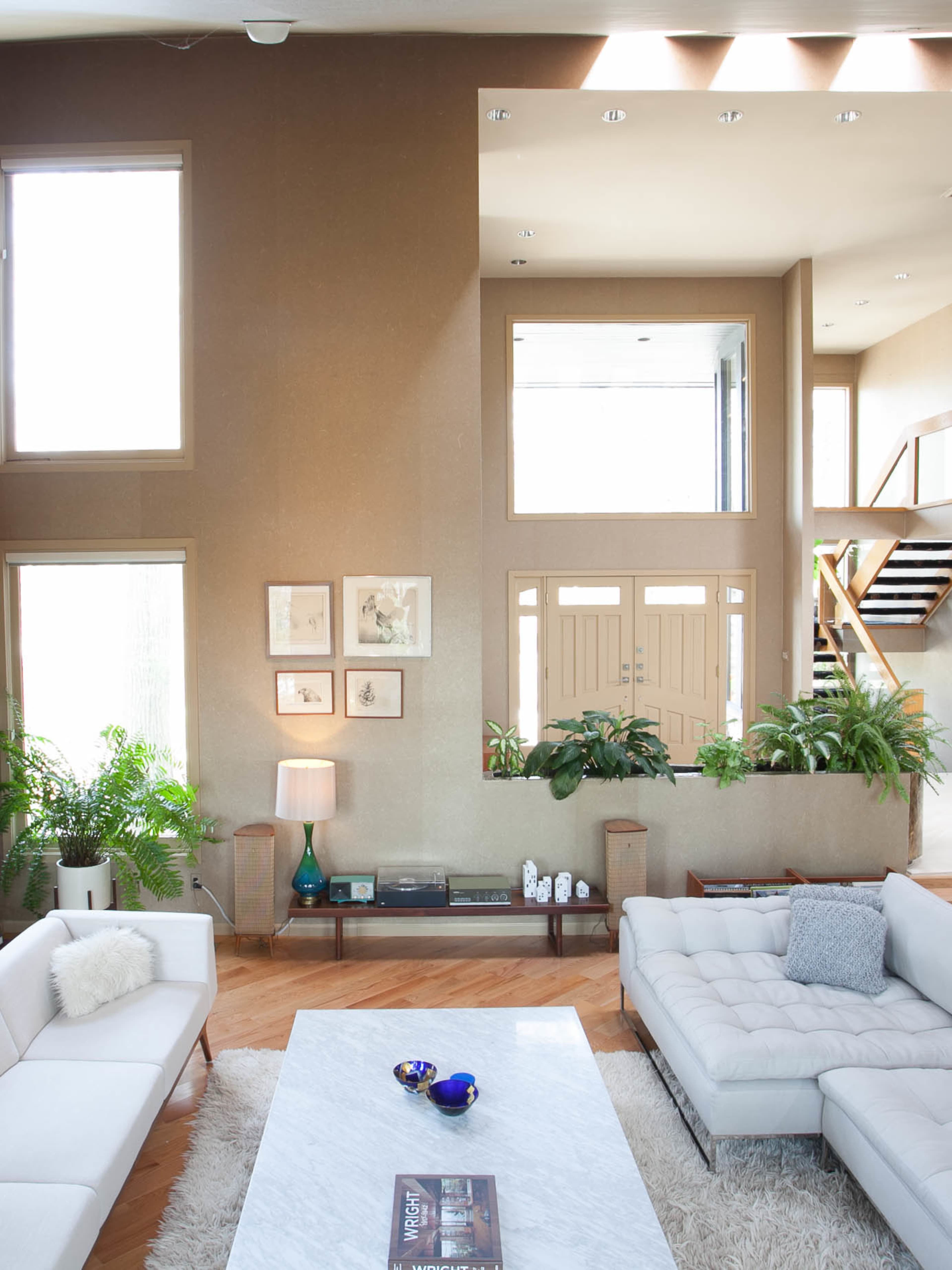 A modern living room features large windows, a marble coffee table, and prominent greenery, with an entrance visible in the background.