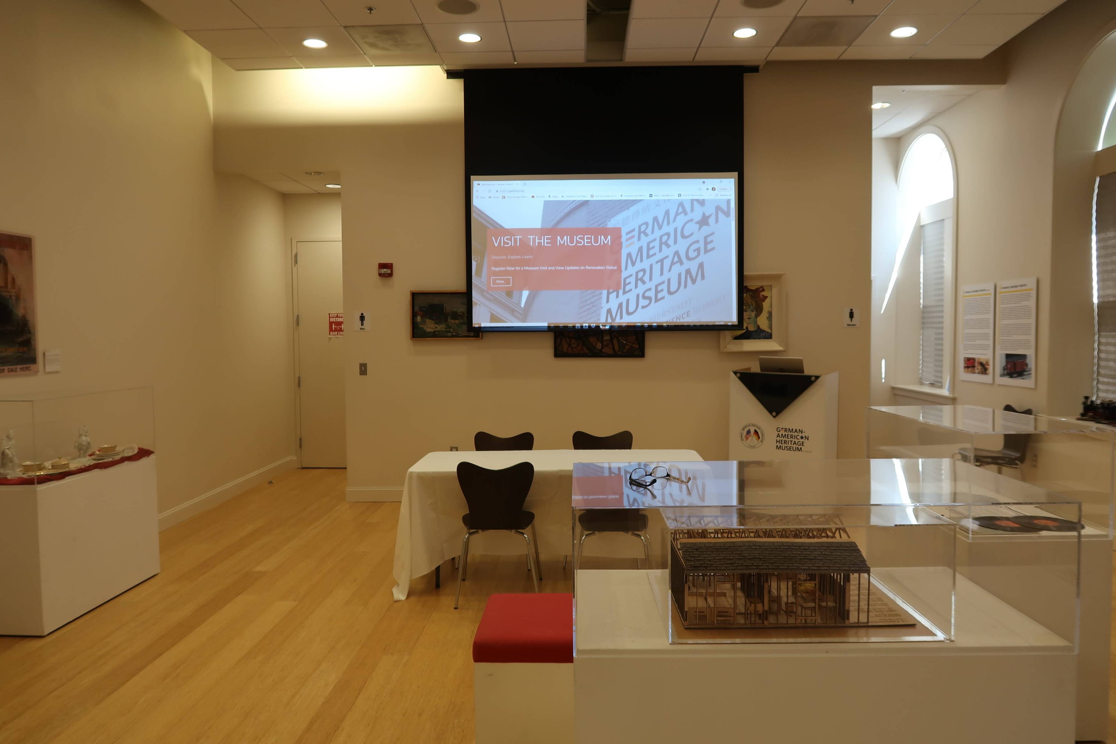 A spacious room in a museum featuring a large screen displaying "Visit the Museum," a table with chairs, and a glass display case showcasing artifacts.