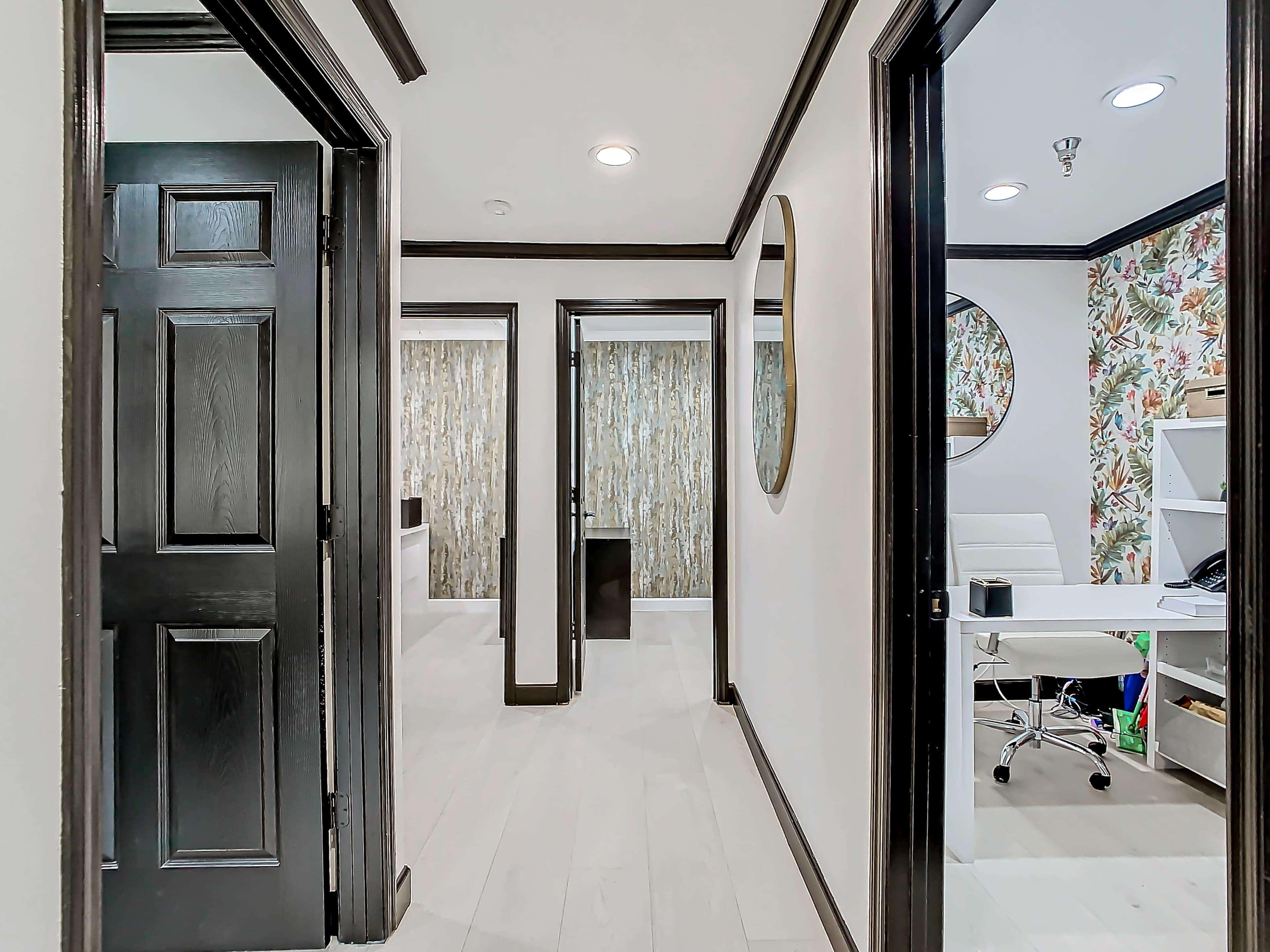 A hallway with black doors leading to rooms, featuring one door on the left and another on the right, with a circular mirror and a desk visible at the end.