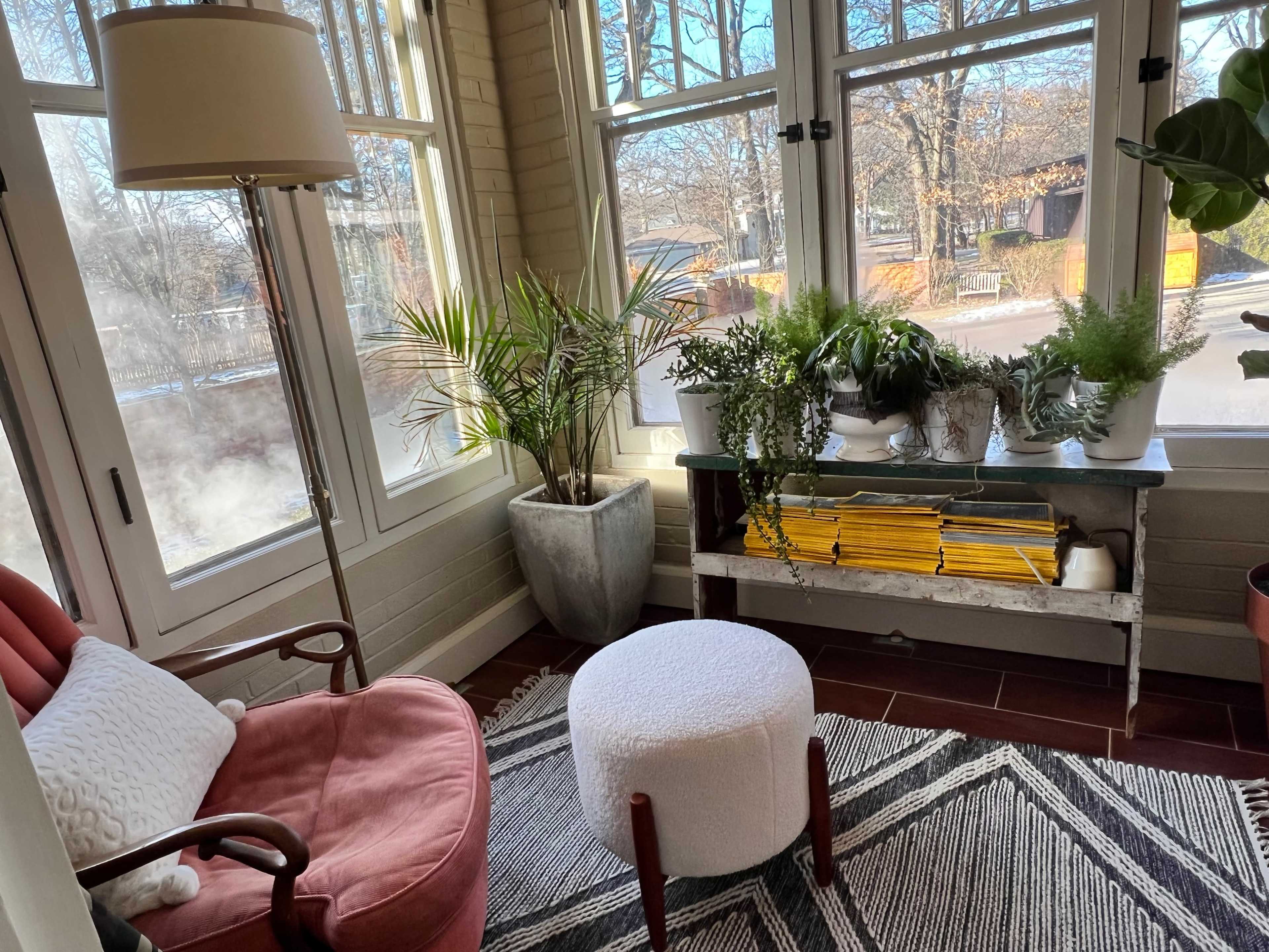 A cozy sunroom features a pink armchair, a white ottoman, potted plants, and a console table with neatly arranged magazines.