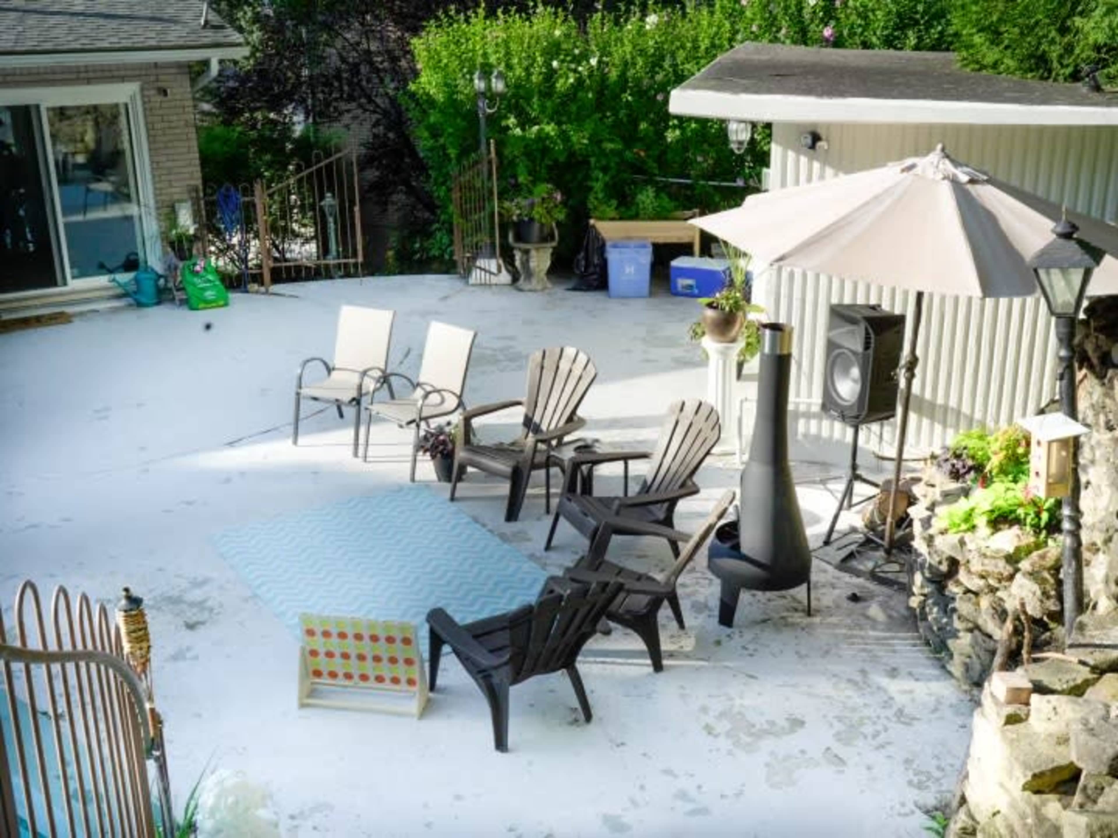 A patio area with several wooden chairs arranged around a blue rug, an umbrella, a speaker, and a stone structure nearby.