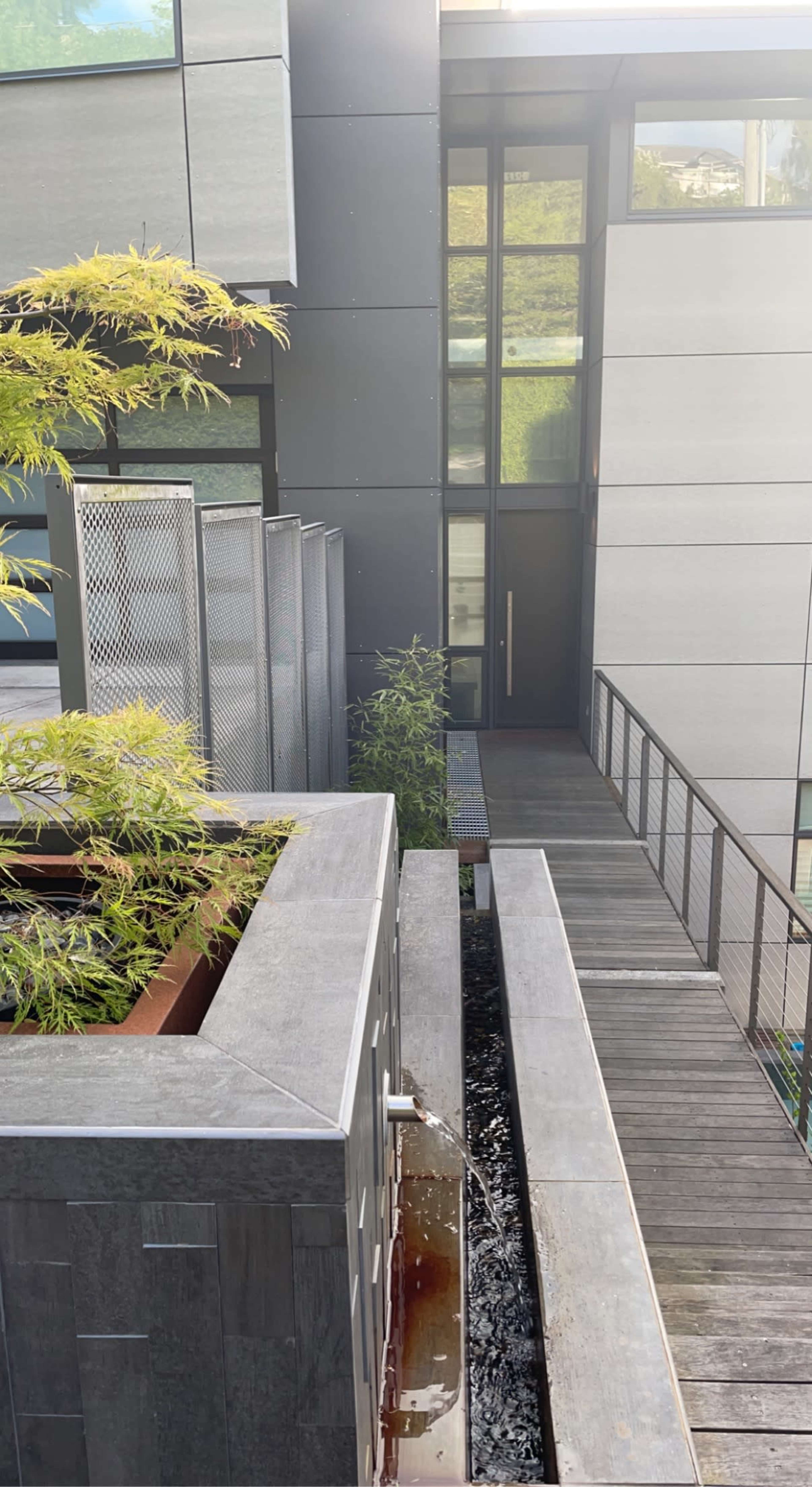 A modern architectural space featuring a wooden walkway, a grey stone planter with a water feature, and large glass windows.