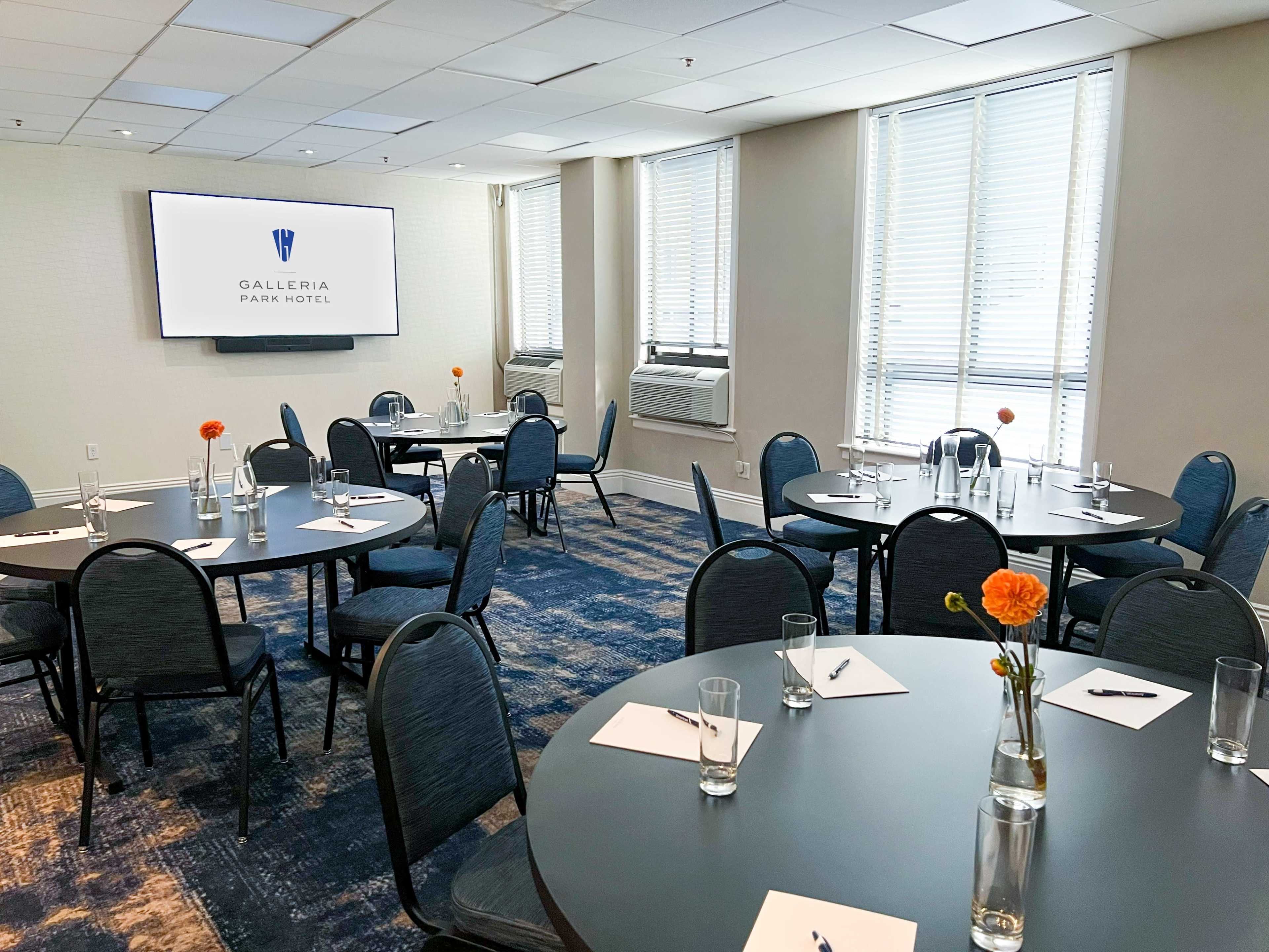 The image shows a hotel meeting room with round tables set for a conference, featuring note pads and glasses of water, and a large screen mounted on the wall.