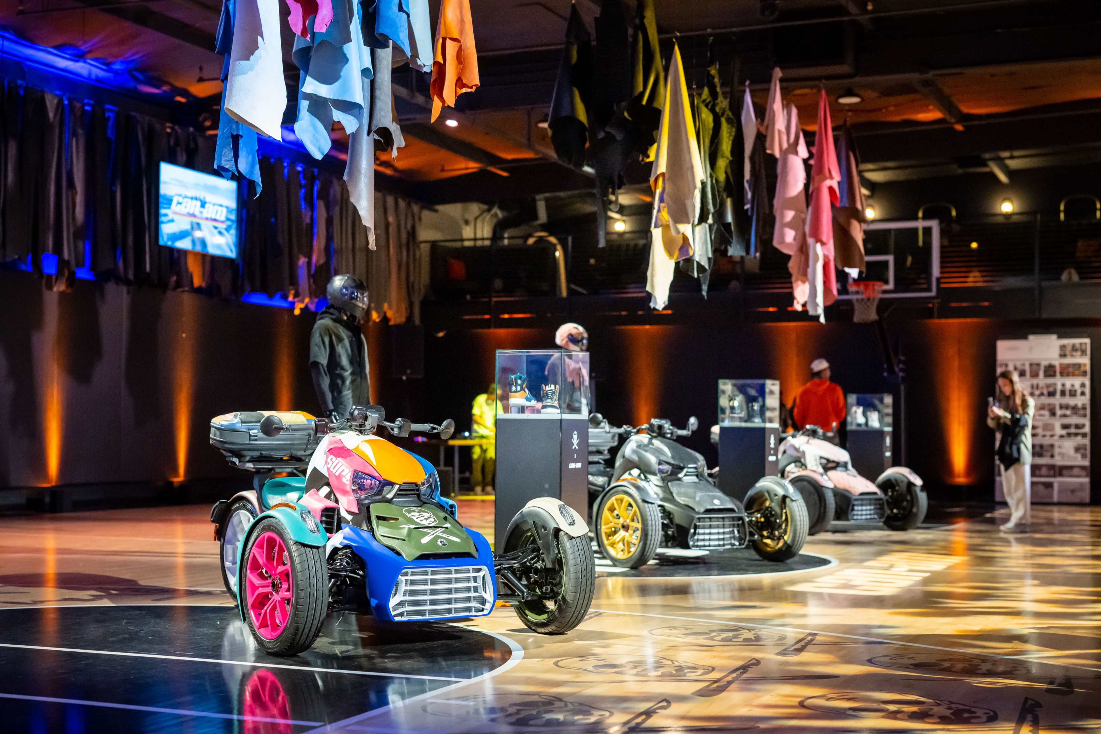 The scene features three colorful quad bikes displayed on a stage with fabric hanging from the ceiling, surrounded by visitors and informational panels.