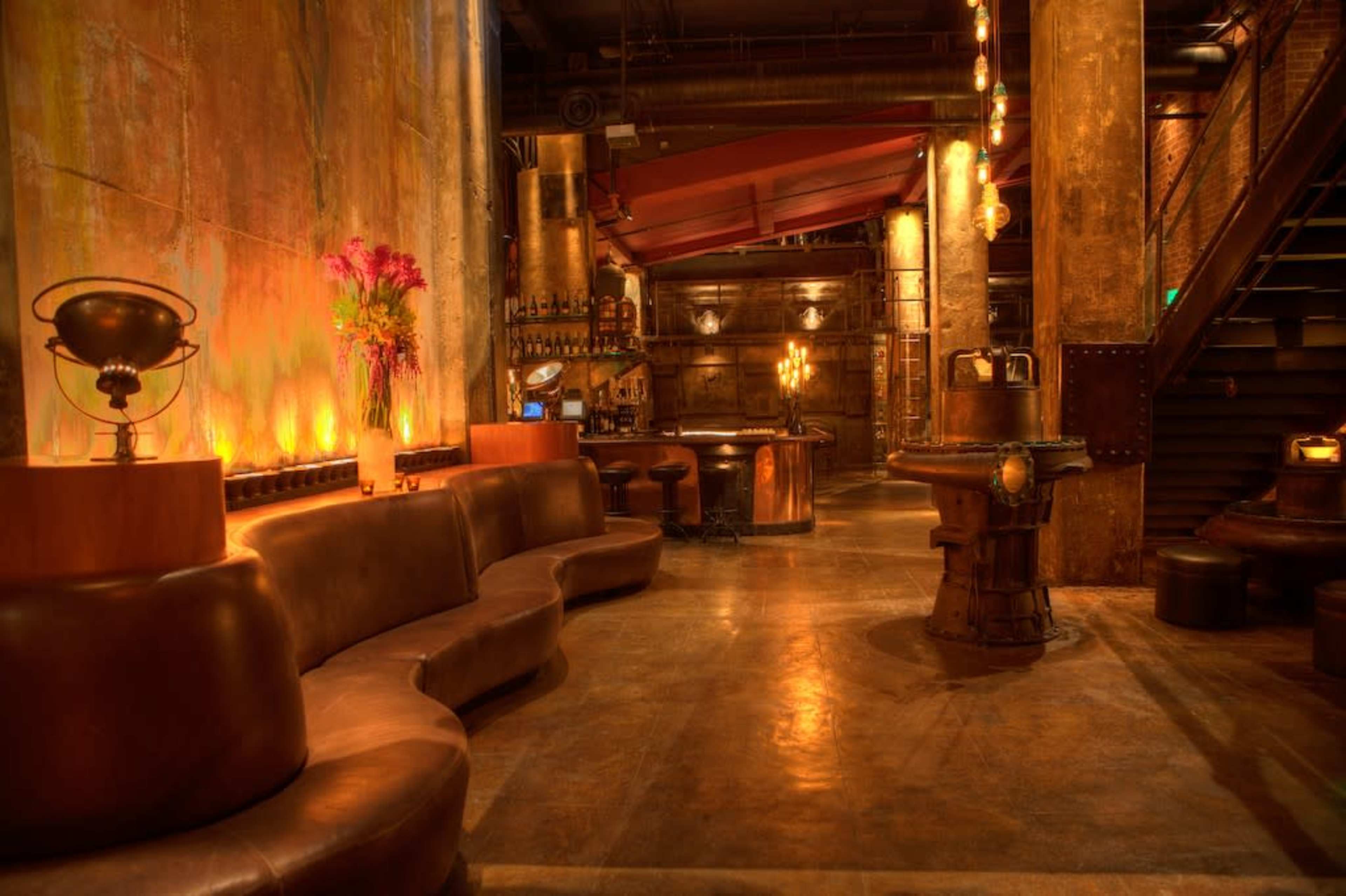The image shows an industrial-style bar area with brown leather seating, illuminated with warm lighting and featuring metallic decor.