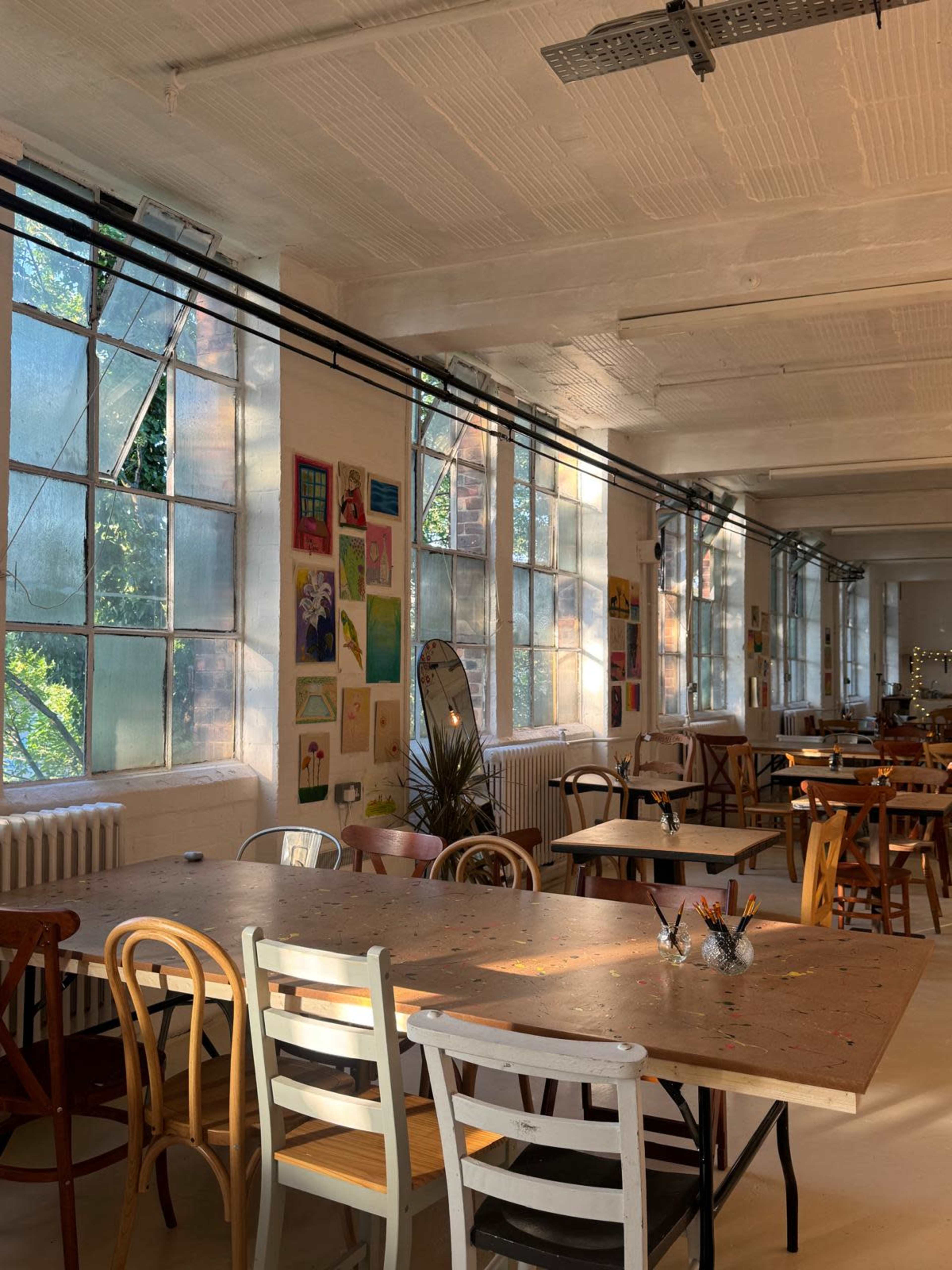 The image shows a bright, spacious room filled with wooden tables and a variety of chairs, illuminated by sunlight streaming through large windows.