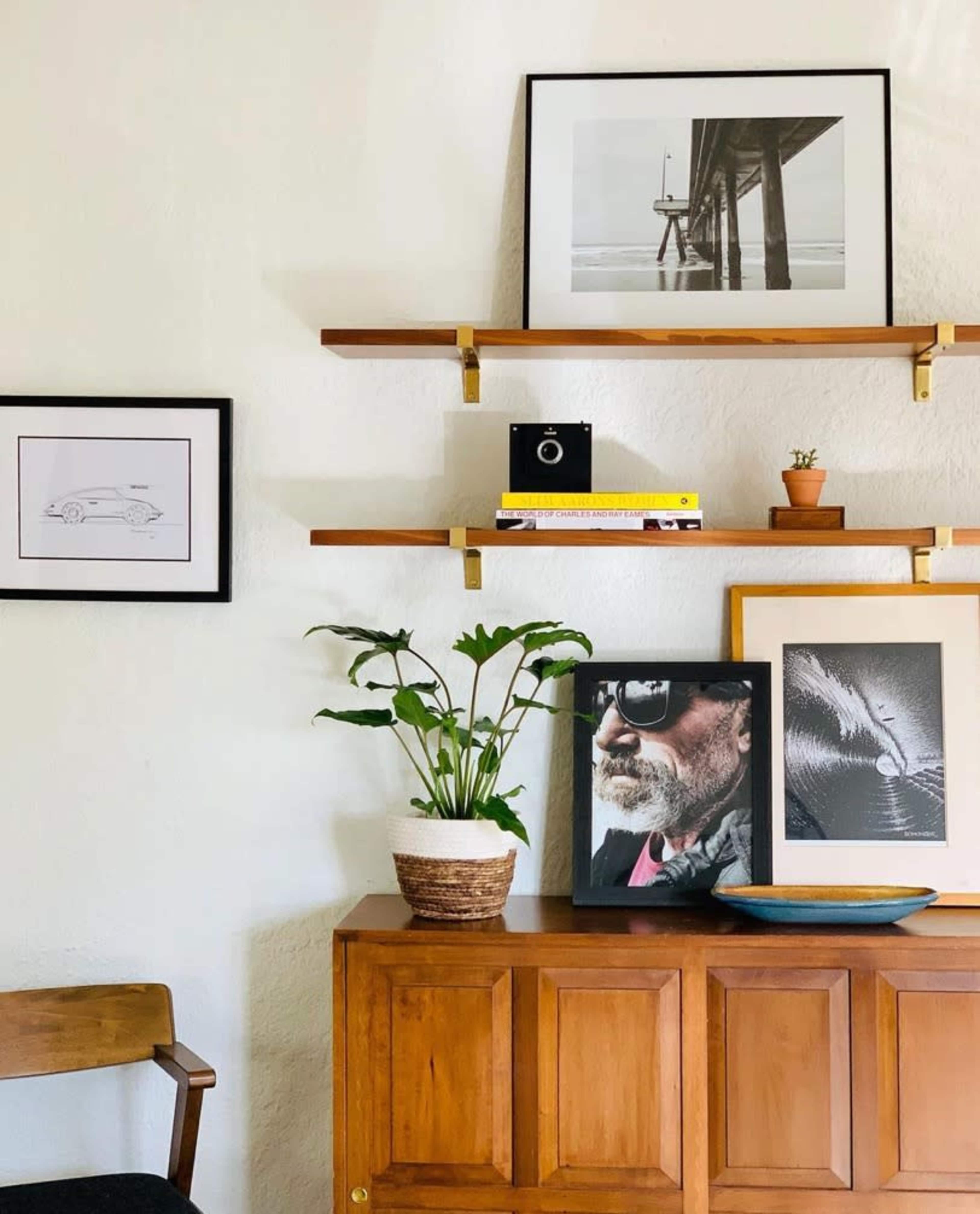 The scene features a wooden cabinet with a collection of framed artwork and decorative items displayed on shelves above it, alongside a potted plant.