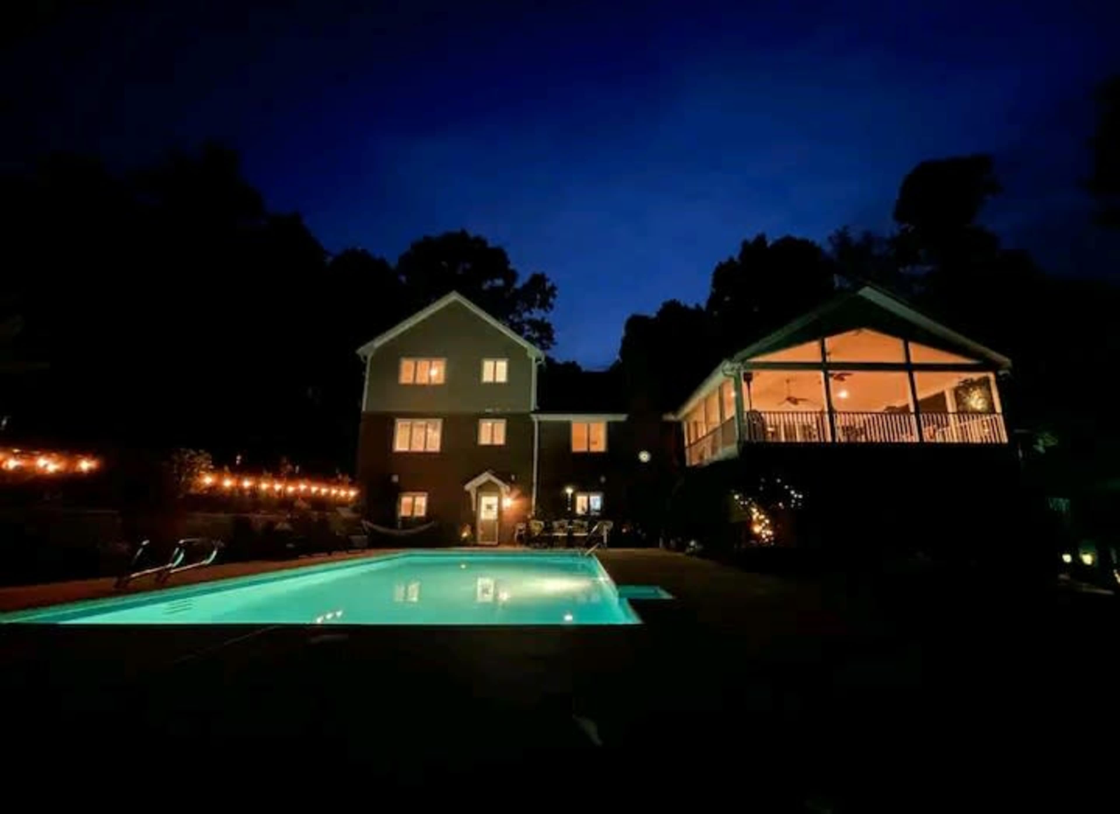 A well-lit pool area is surrounded by a two-story house and a covered porch at dusk.