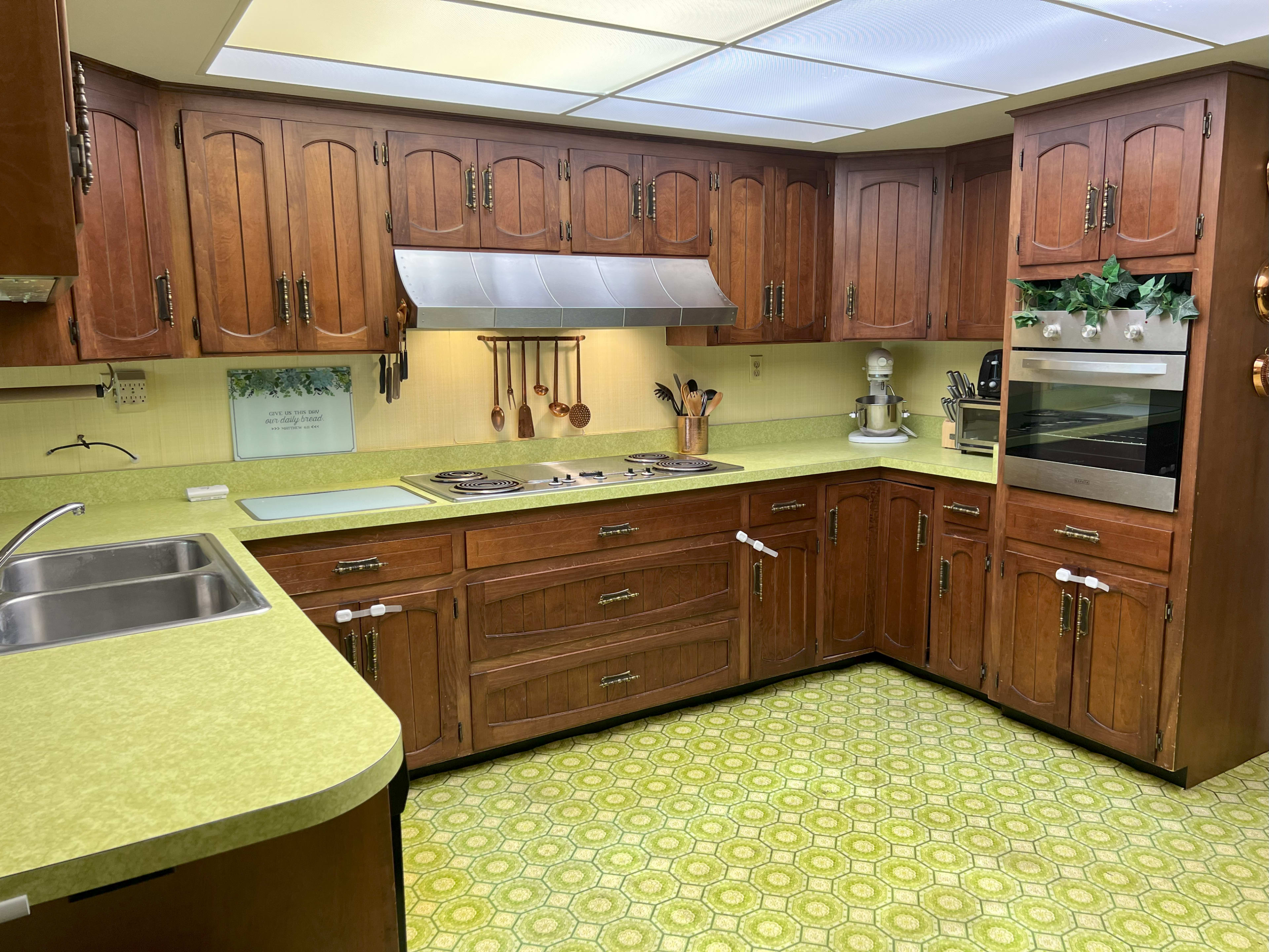 The image shows a retro-style kitchen with wooden cabinetry, a green countertop, and patterned linoleum flooring.
