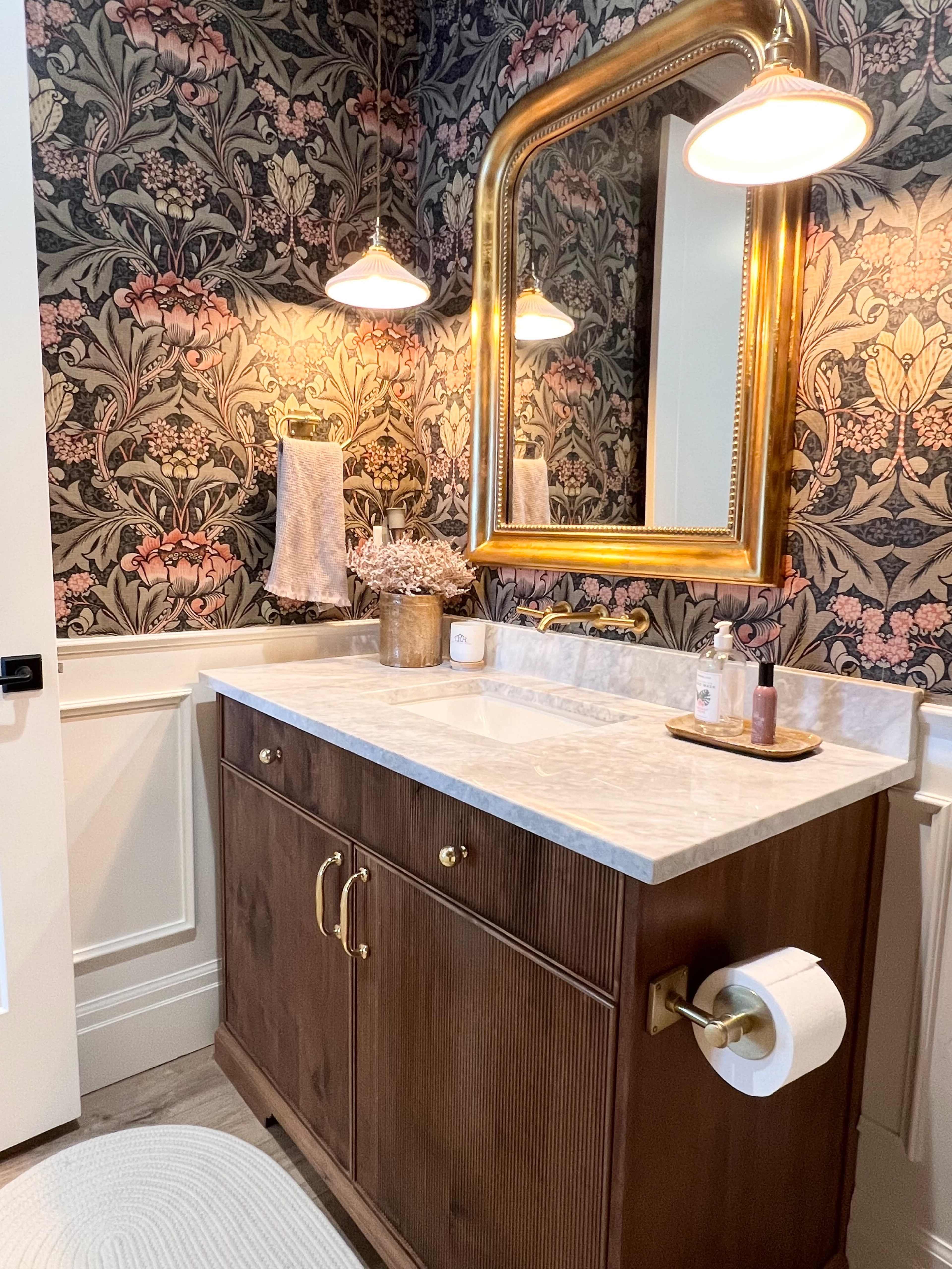 A bathroom with floral wallpaper, a wooden vanity with gold hardware, and a large mirror framed in gold.