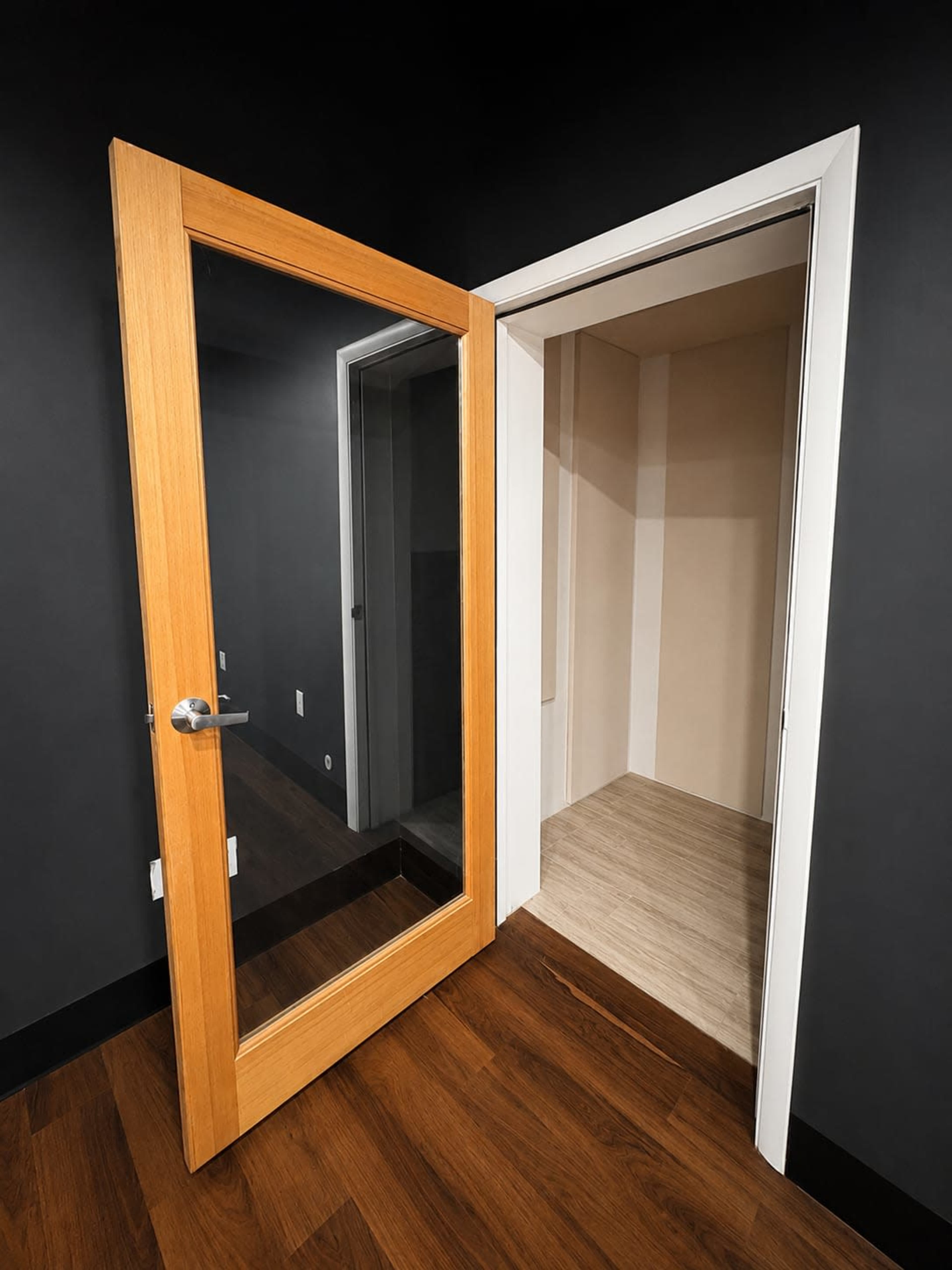 A wooden door with a glass panel is open, revealing a small, empty room adjacent to a dark wall and hardwood floor.