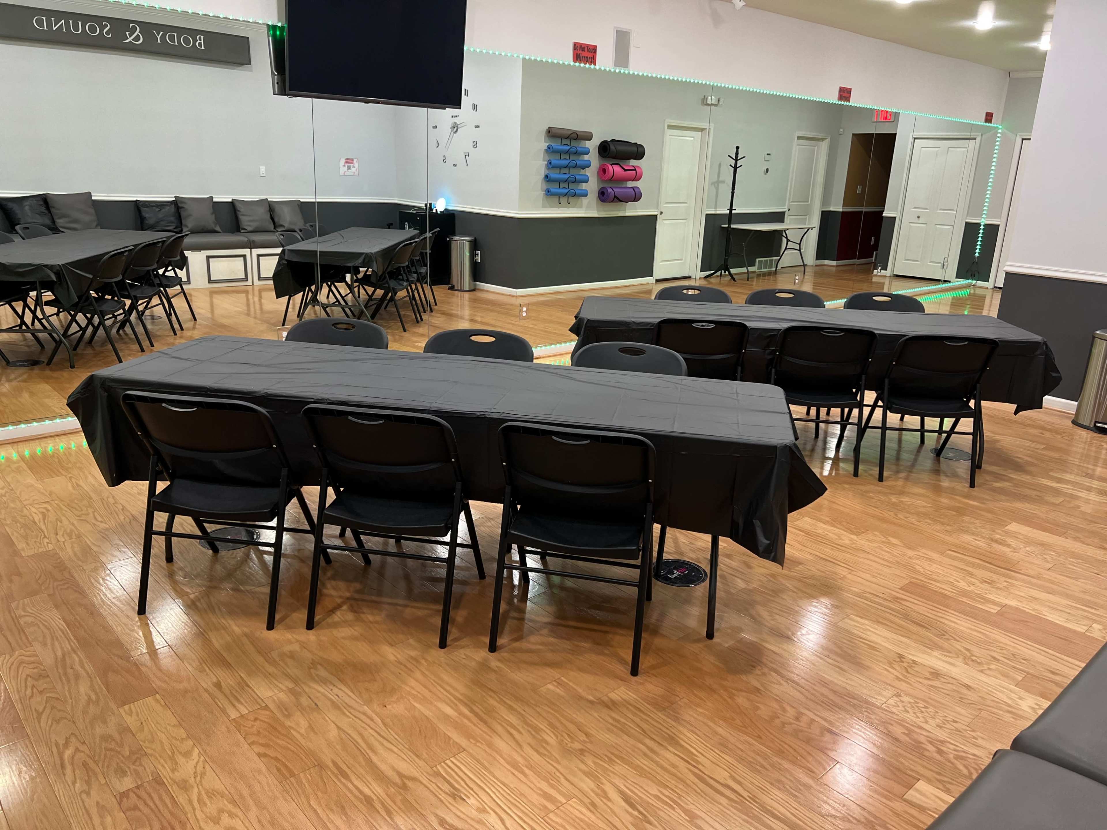 A room with a polished wooden floor features several black-covered tables and chairs arranged for an event, with mirrors on one wall and exercise mats stacked in the background.