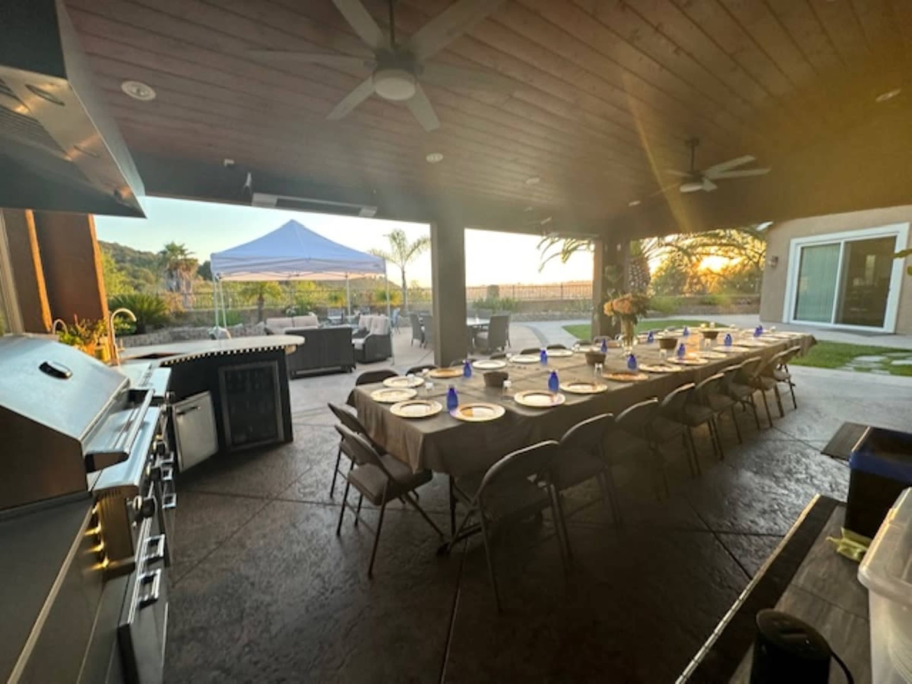 A spacious outdoor dining area features a long table set with blue glasses and white plates, alongside a grill and seating area under a covered patio.