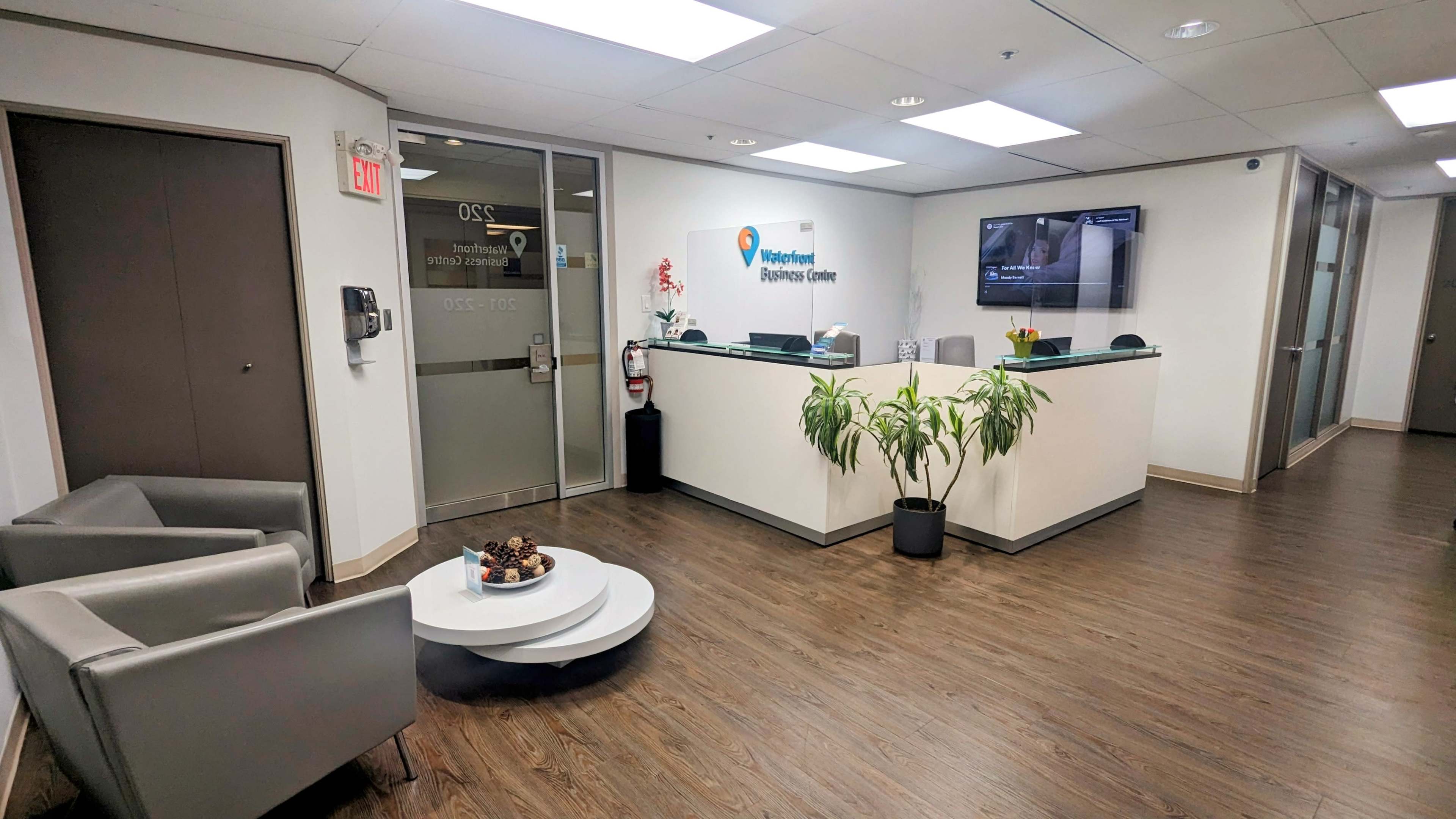 A modern reception area with a front desk, two gray chairs, a coffee table with a decorative bowl, and potted plants.
