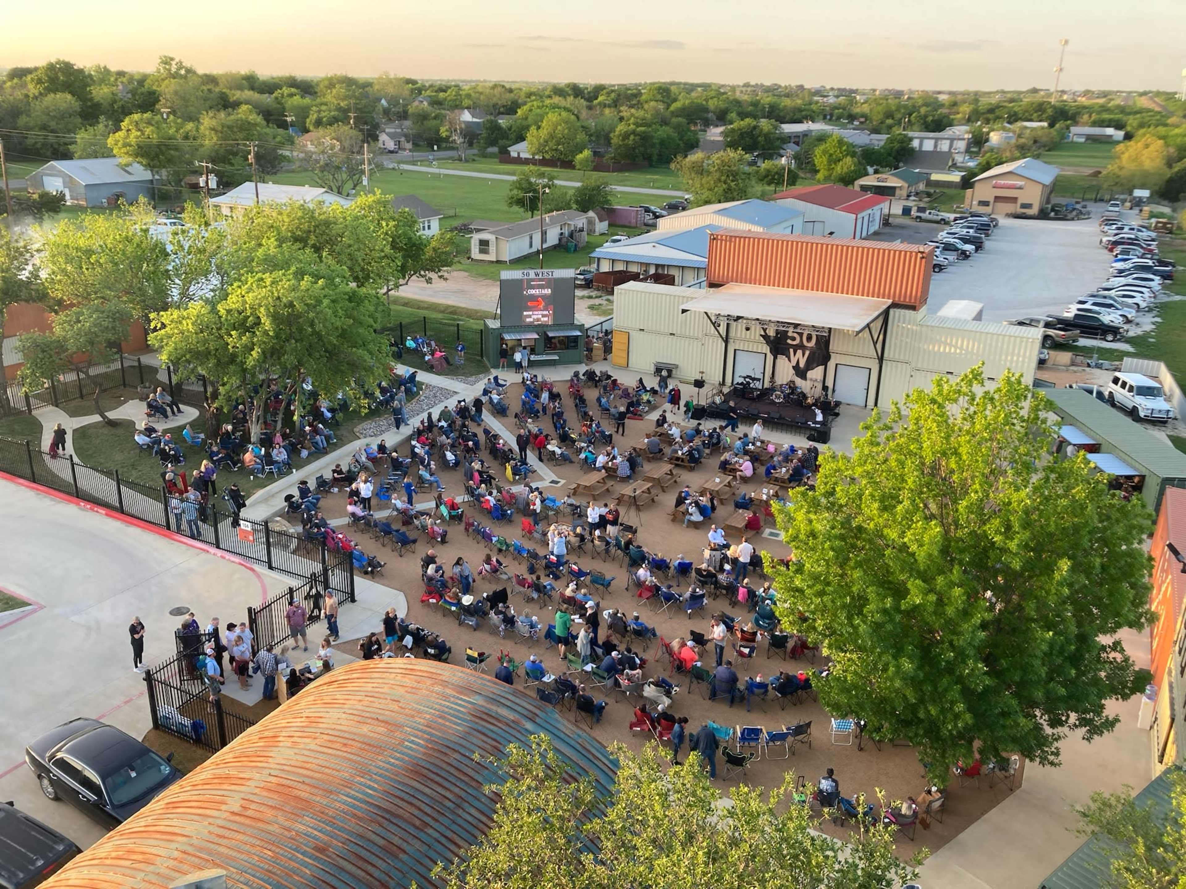 A large outdoor audience sits in folding chairs, arranged in rows, facing a stage at a community event area.