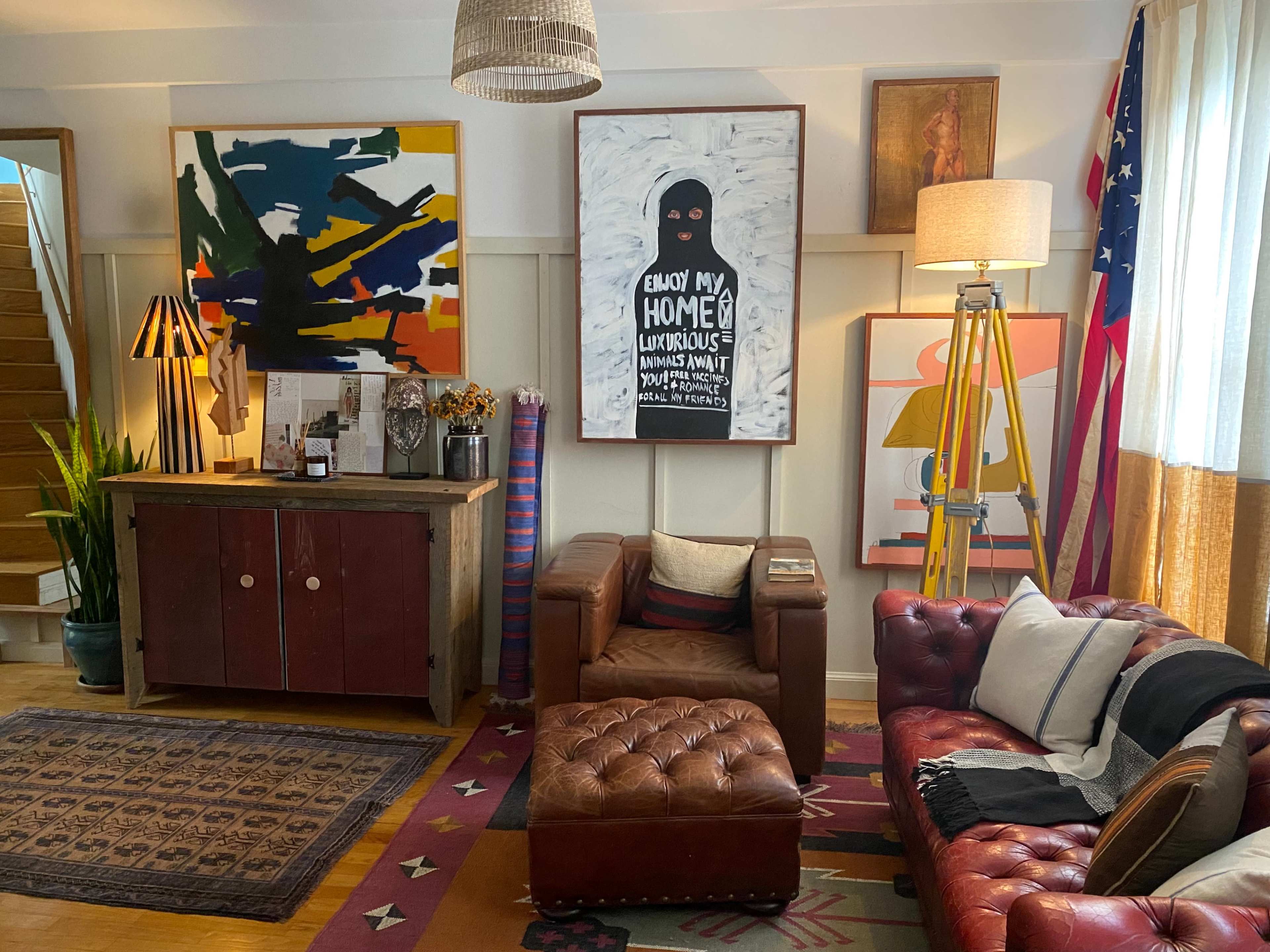 The image shows a cozy living room featuring a brown leather sofa, a wooden sideboard, colorful artwork on the walls, and an American flag in the corner.