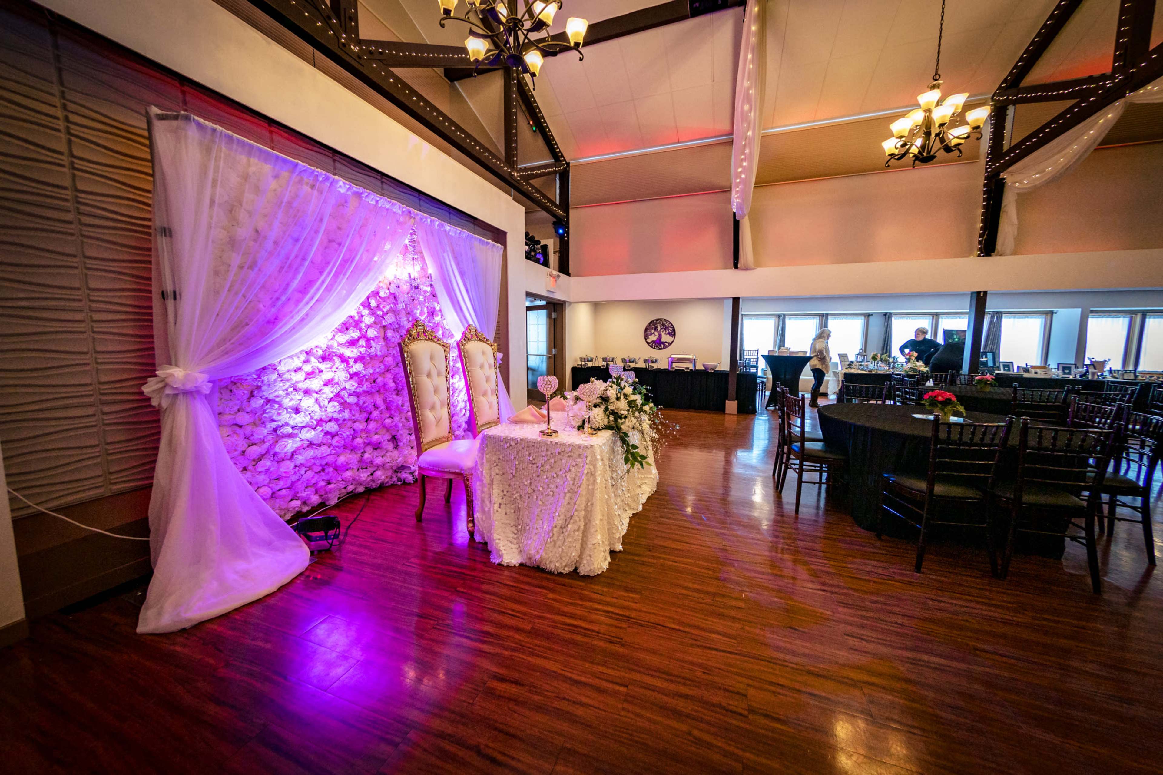 A decorated wedding reception area features a floral backdrop, a head table adorned with a white tablecloth, and dark wooden chairs in a spacious venue.