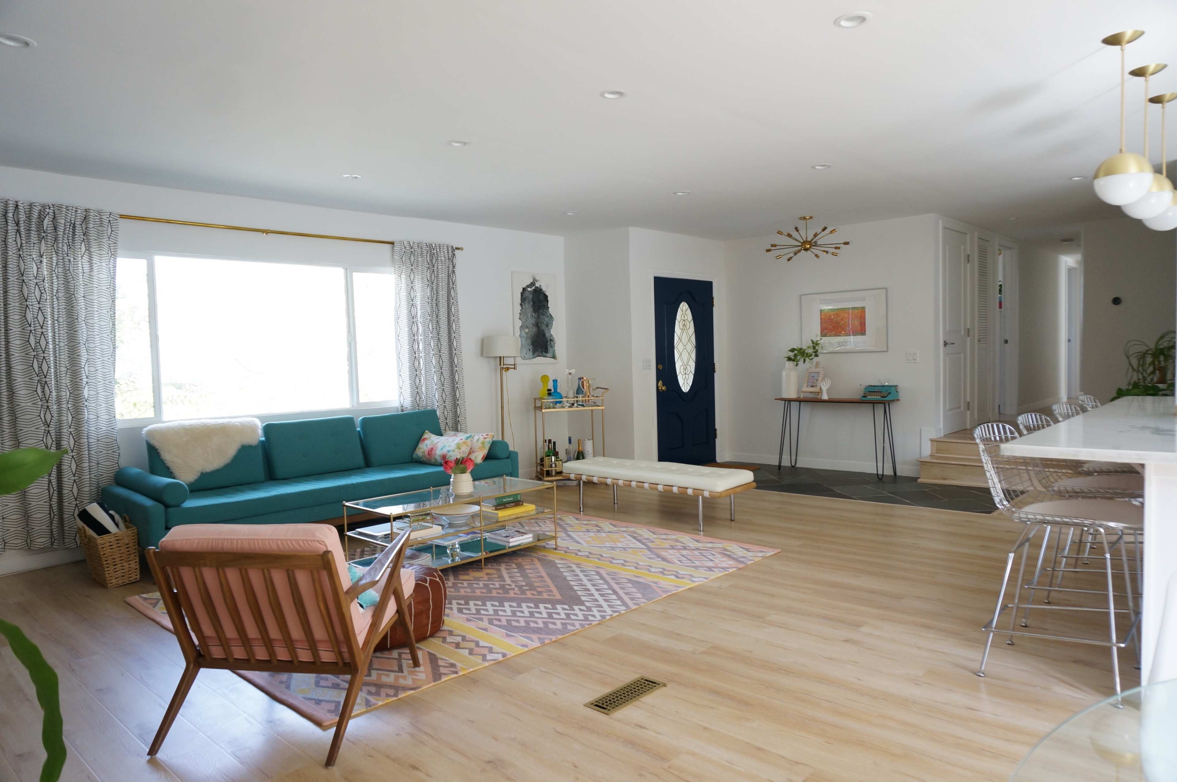 A modern living room features a teal sofa, a patterned area rug, a coffee table, and a view of a large window with natural light.