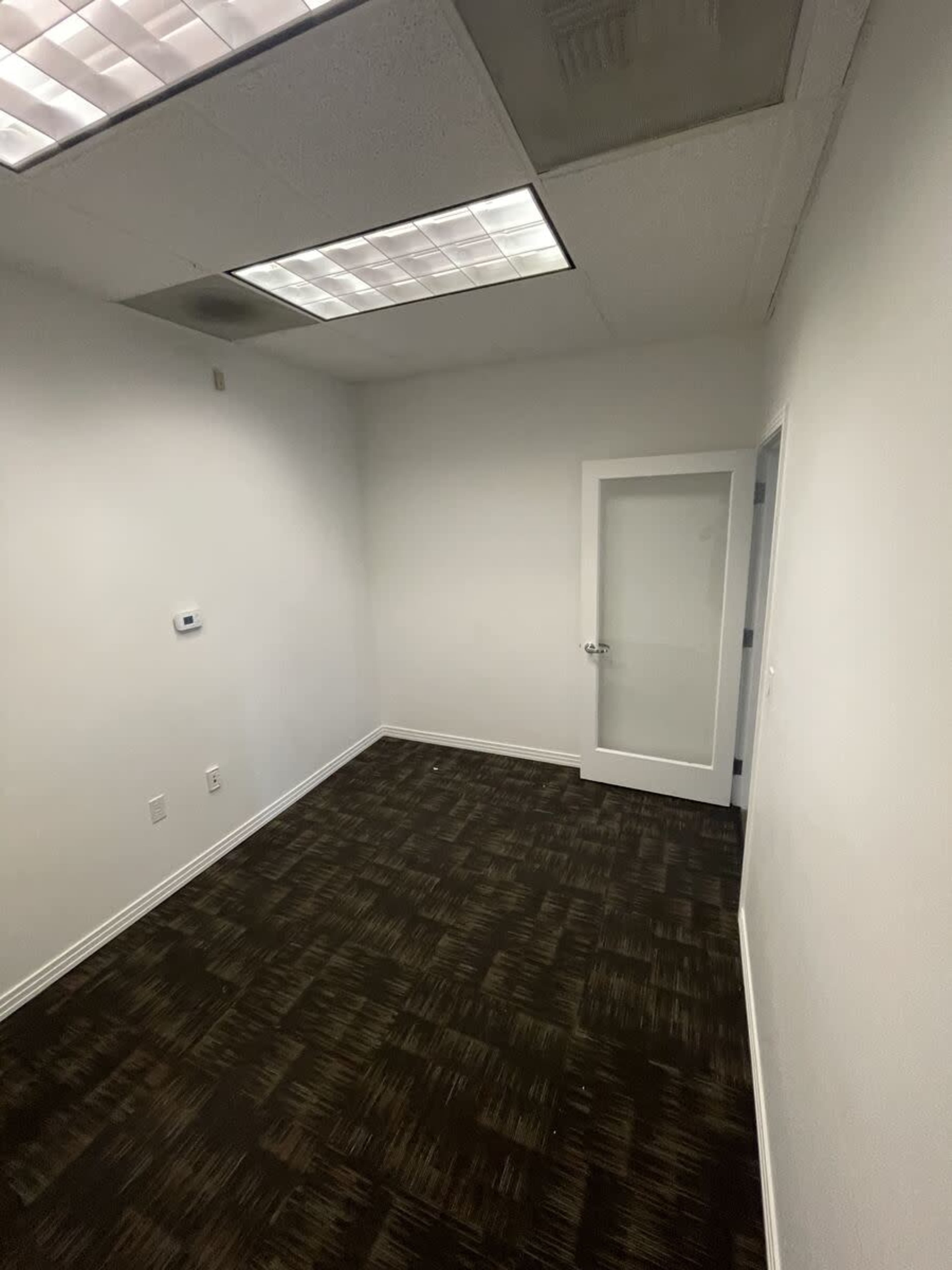The image shows a vacant office space with white walls, a door, and a carpeted floor.