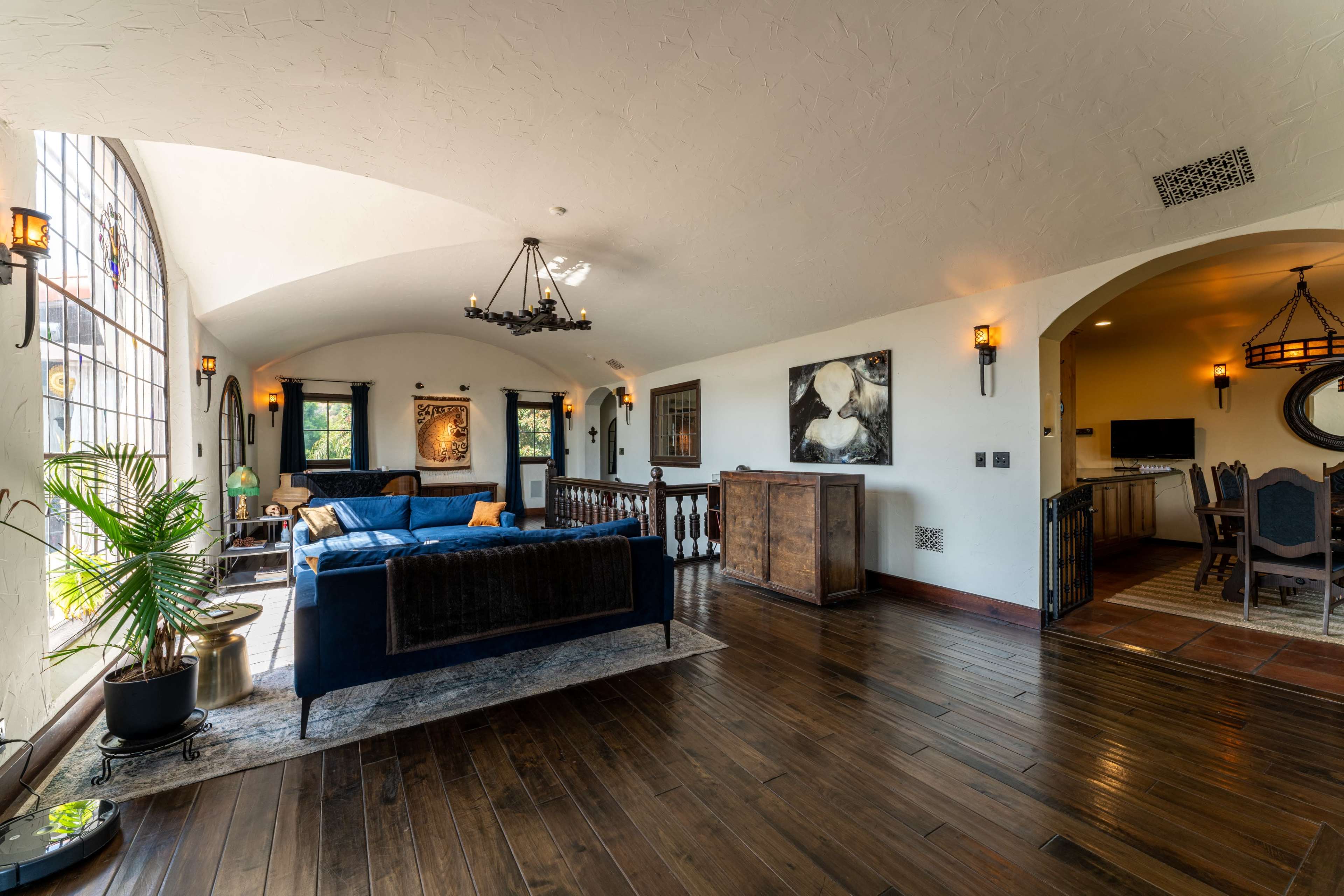 The image depicts a spacious living area with wooden floors, large arched windows, and a mix of modern and traditional furniture.