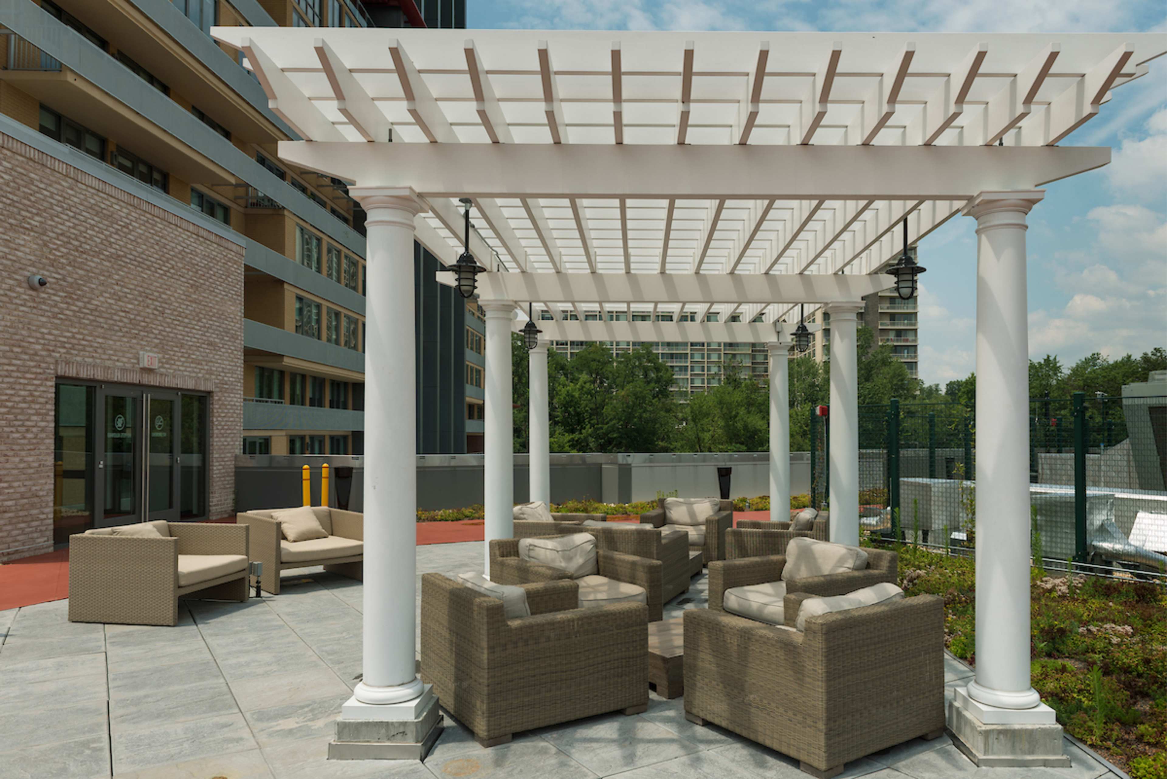 A pergola supported by white columns provides shaded seating with wicker chairs and cushions in an outdoor area next to a building.