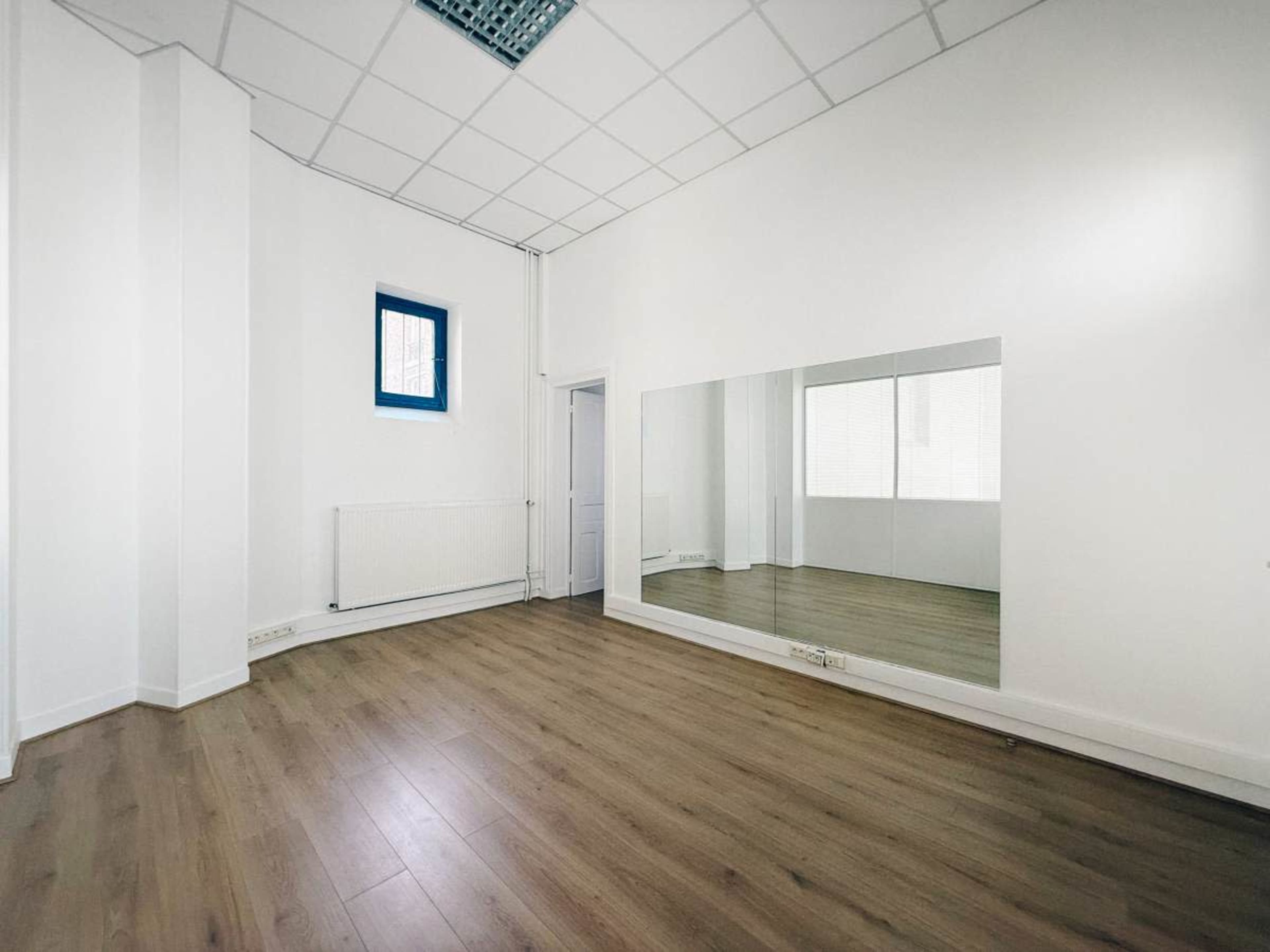 The image shows an empty room with wooden flooring, a single window, and a large mirror on one wall.