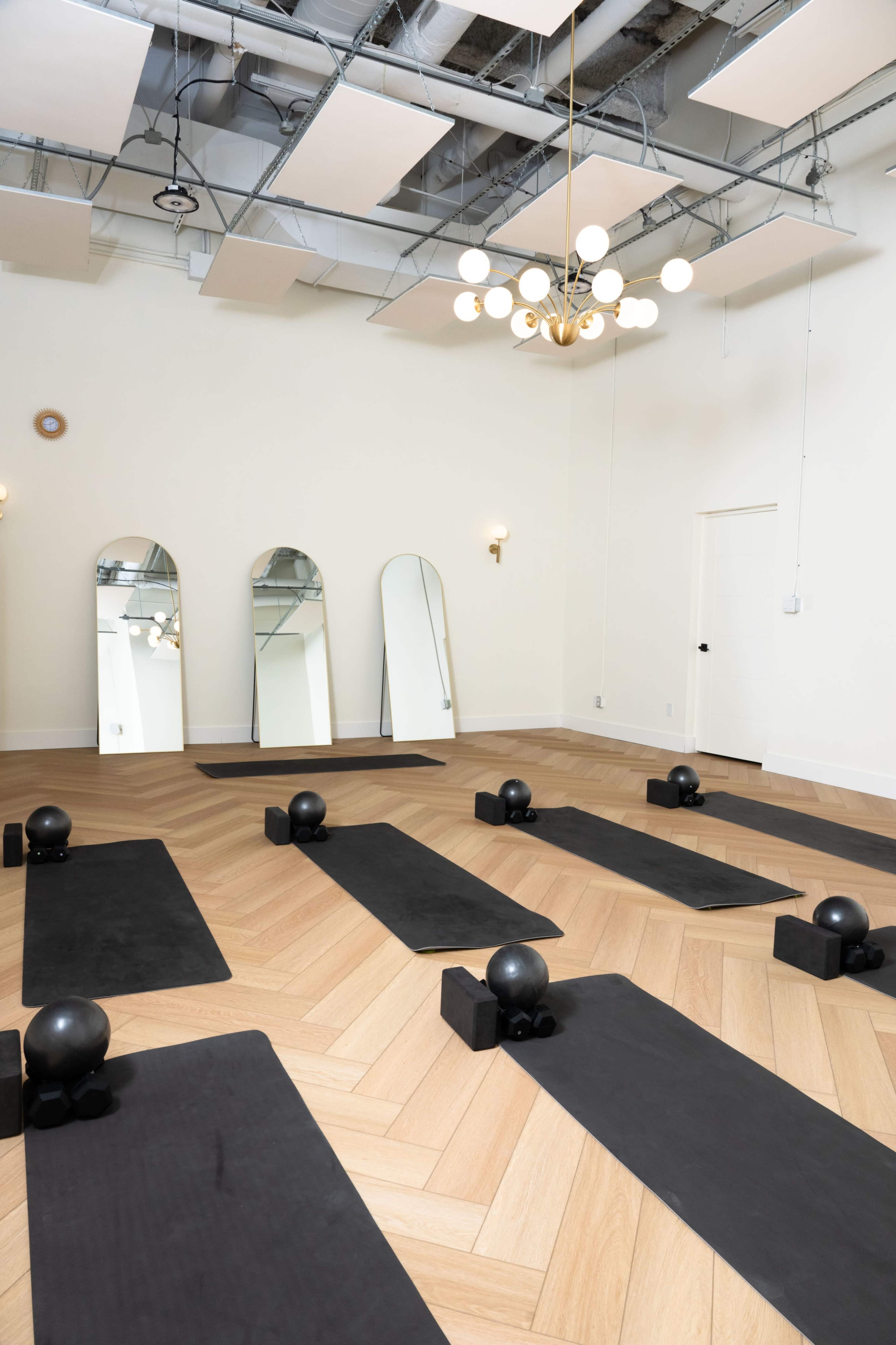 The image shows a fitness studio featuring several exercise mats arranged on a wooden floor, with black weights positioned on each mat and three large mirrors on the wall.