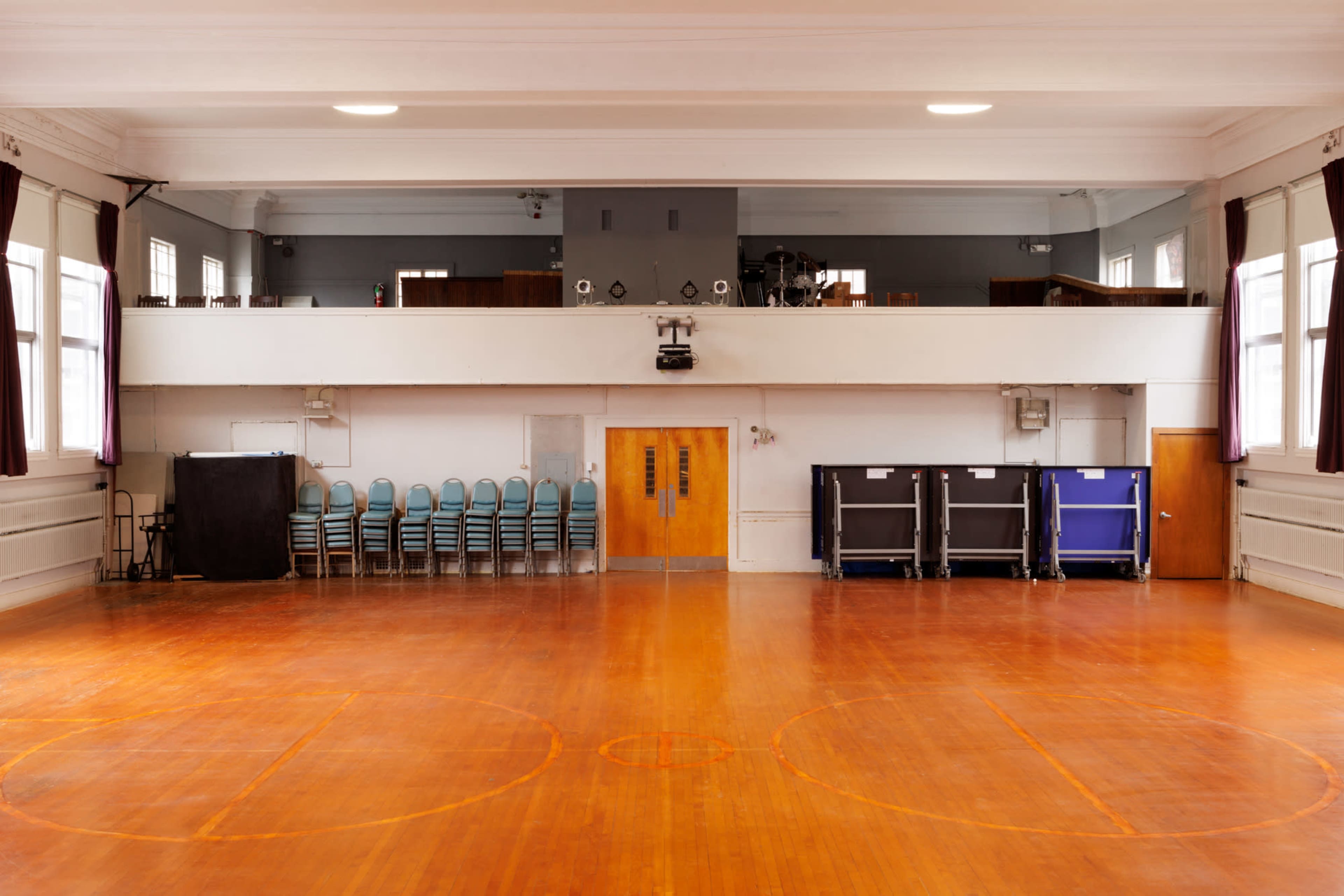 An empty gymnasium with a polished wooden floor, chairs stacked against the wall, and a stage area in the background.