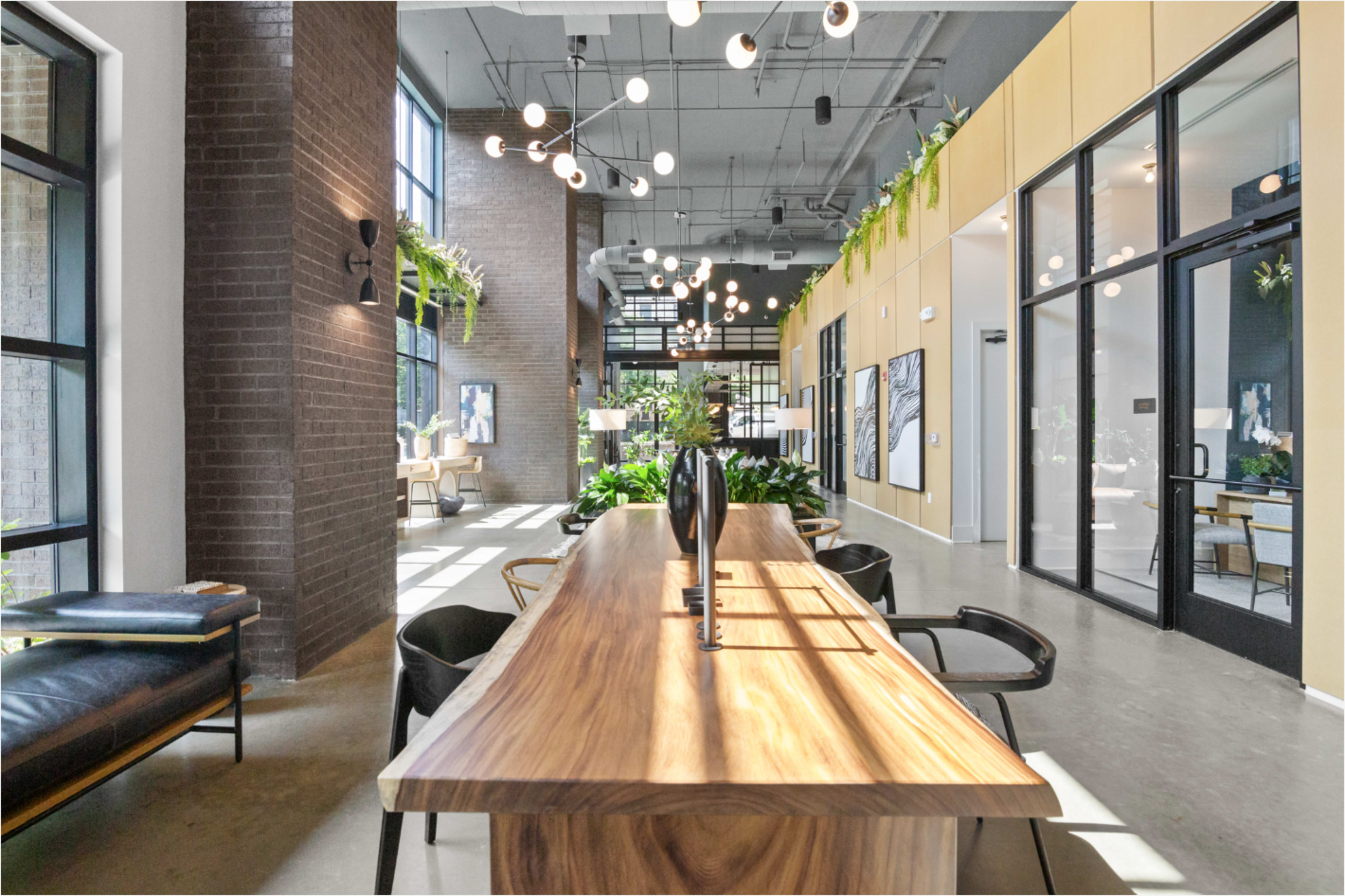 The image showcases a modern lobby area featuring a long wooden table, vibrant indoor plants, and large windows that allow natural light to fill the space.