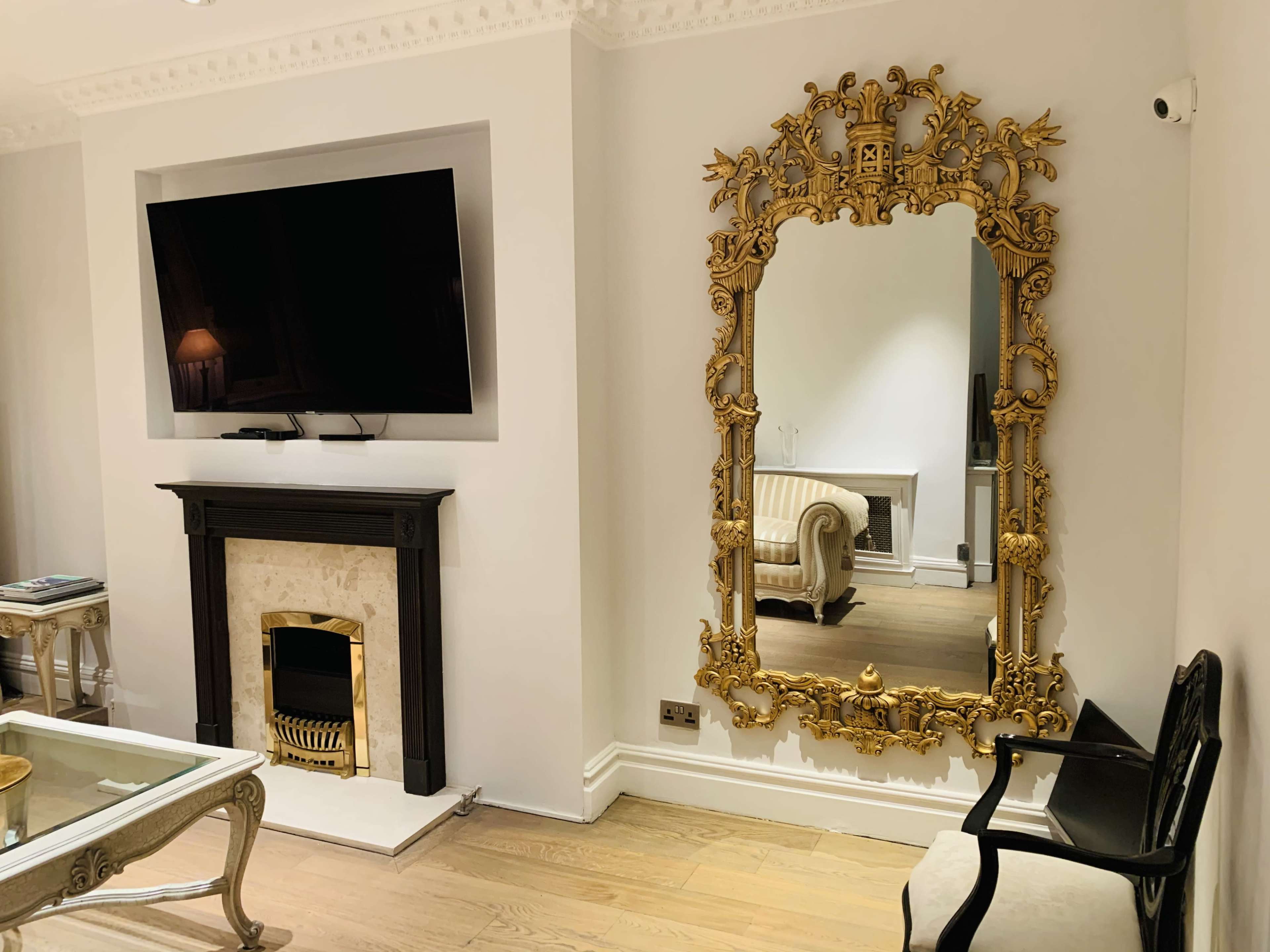 The room features a large ornate gold mirror opposite a flat-screen television mounted on a wall, with a marble fireplace and a beige sofa in the background.