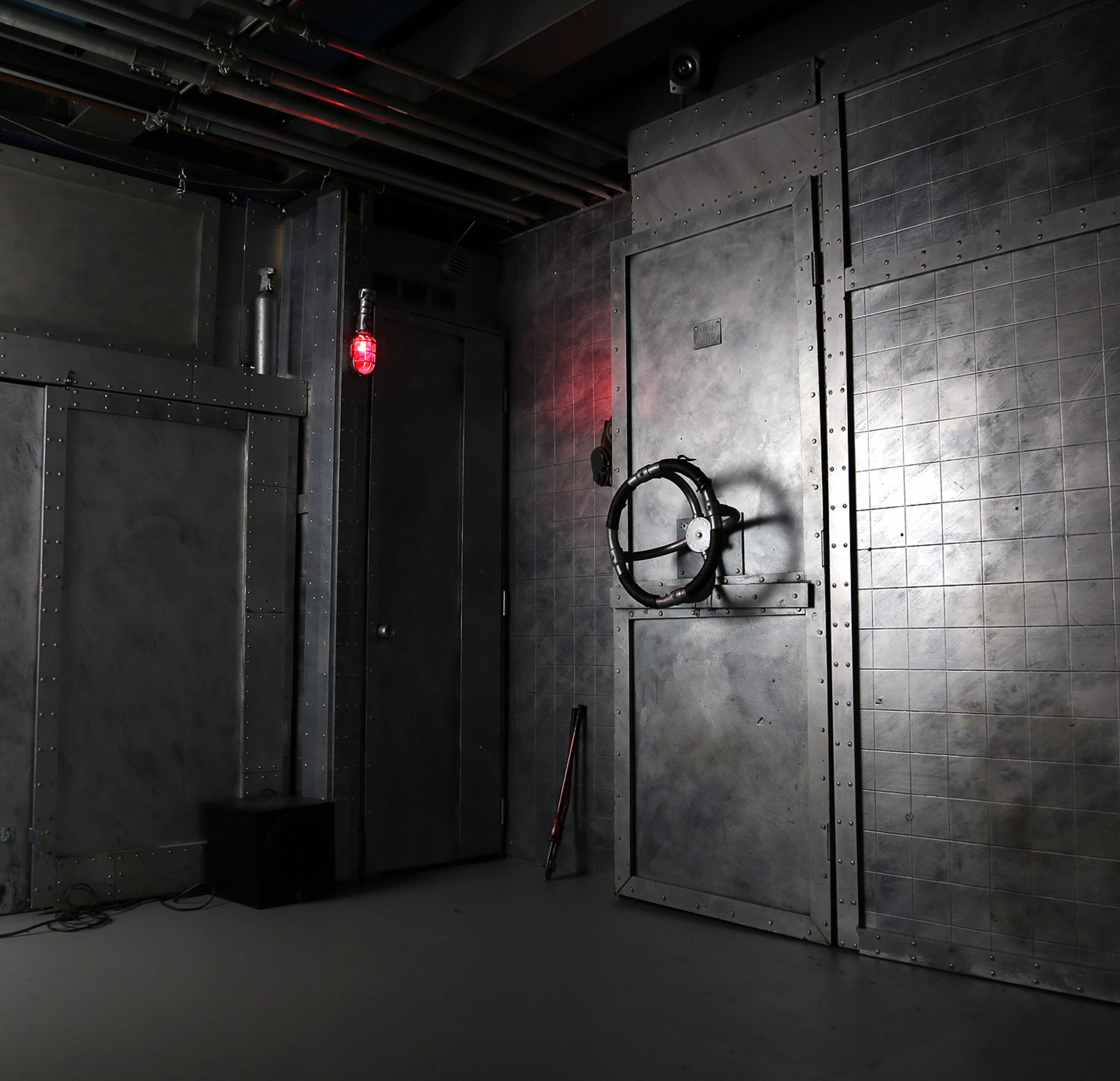 A dimly lit room with metallic walls, featuring a large door with a wheel and a red light in the upper corner.