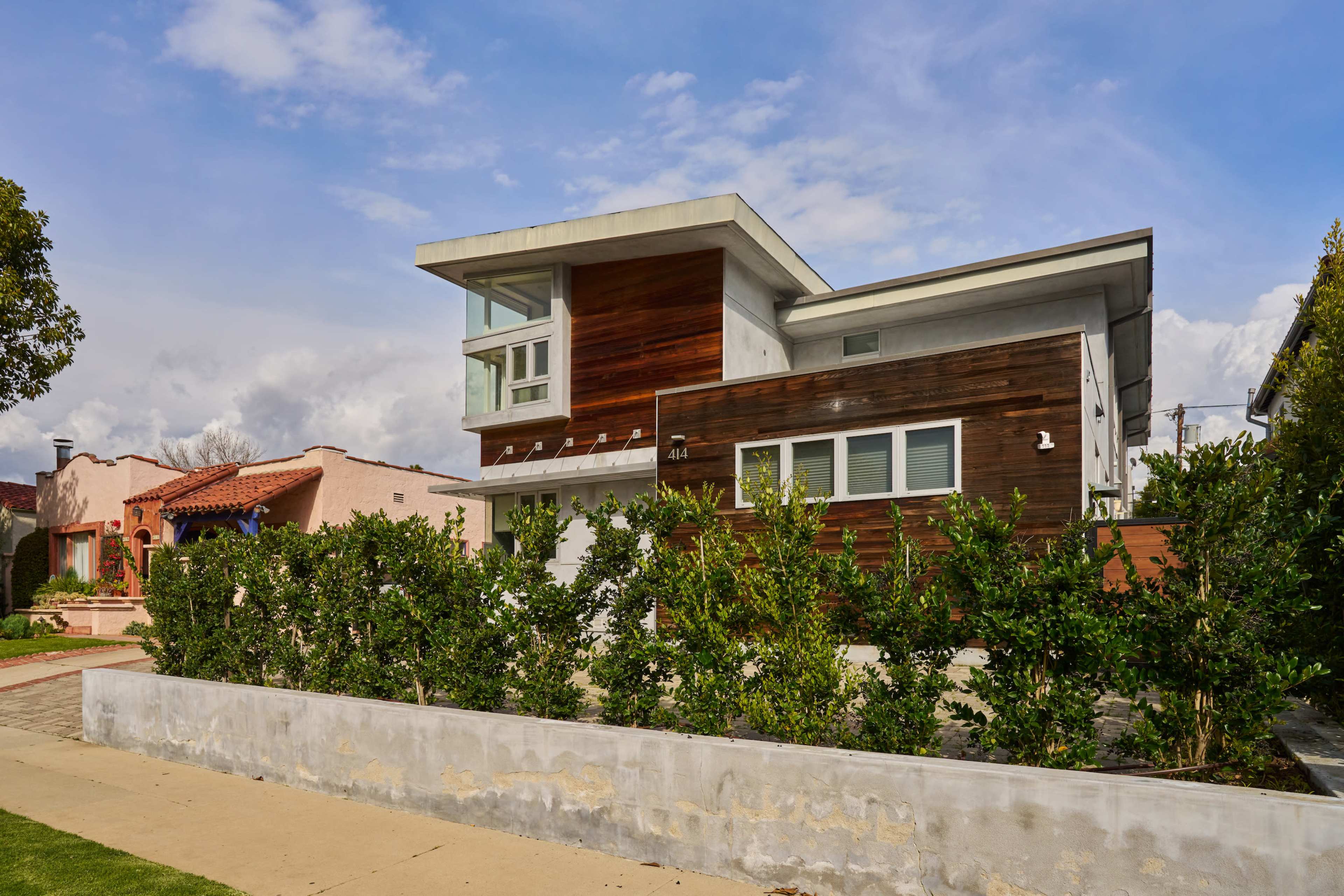 The image shows a modern two-story house with a mix of wood and concrete exterior, surrounded by a low hedge and a concrete pathway.