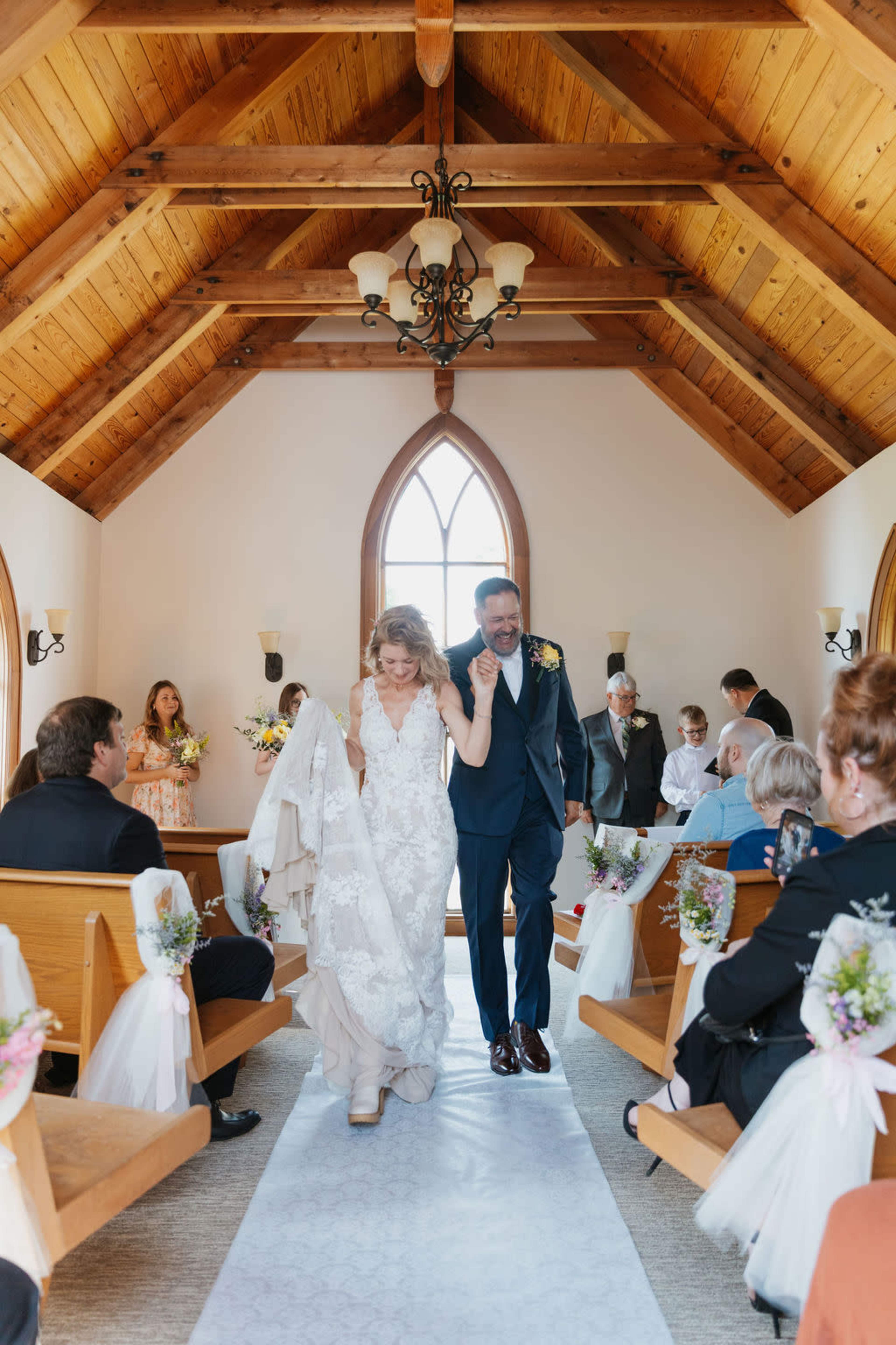 A bride and groom walk down the aisle in a wooden chapel, surrounded by seated guests and floral decorations along the pews.