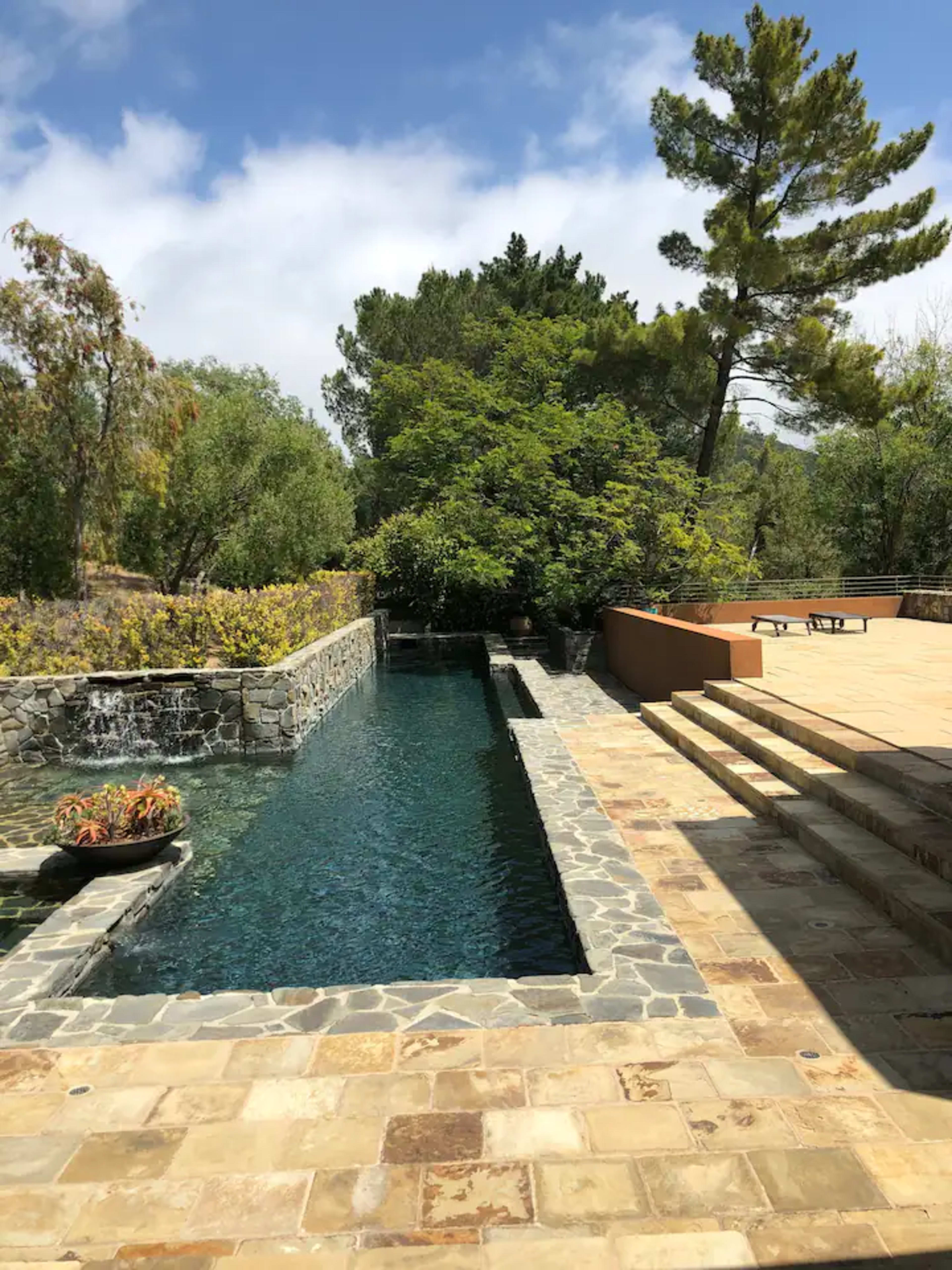 The image shows a stone patio with steps leading down to a narrow pool of water bordered by greenery and a small waterfall.