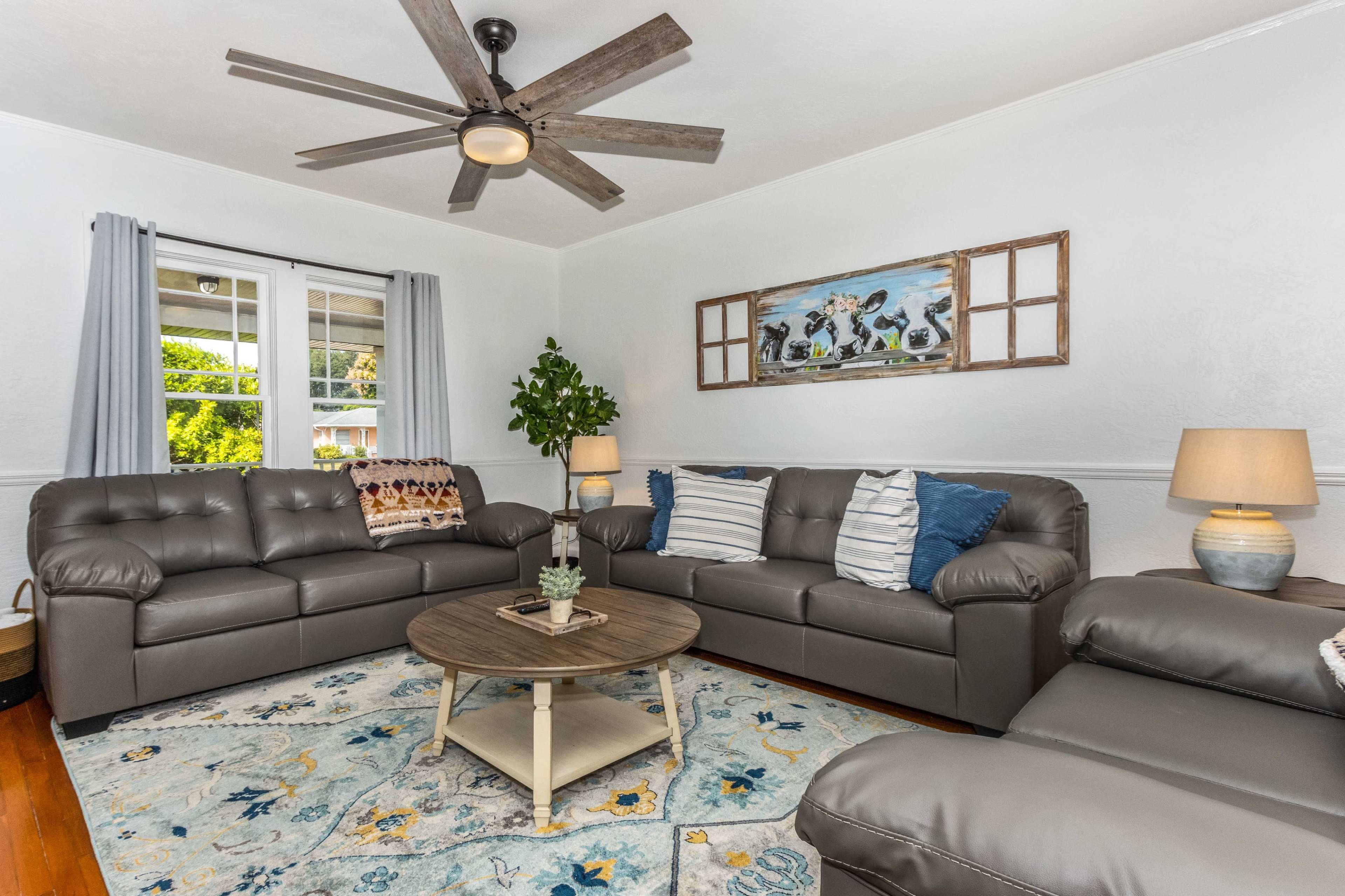 A living room features gray leather sofas, a round wooden coffee table, and a decorative blue and yellow area rug under a ceiling fan.