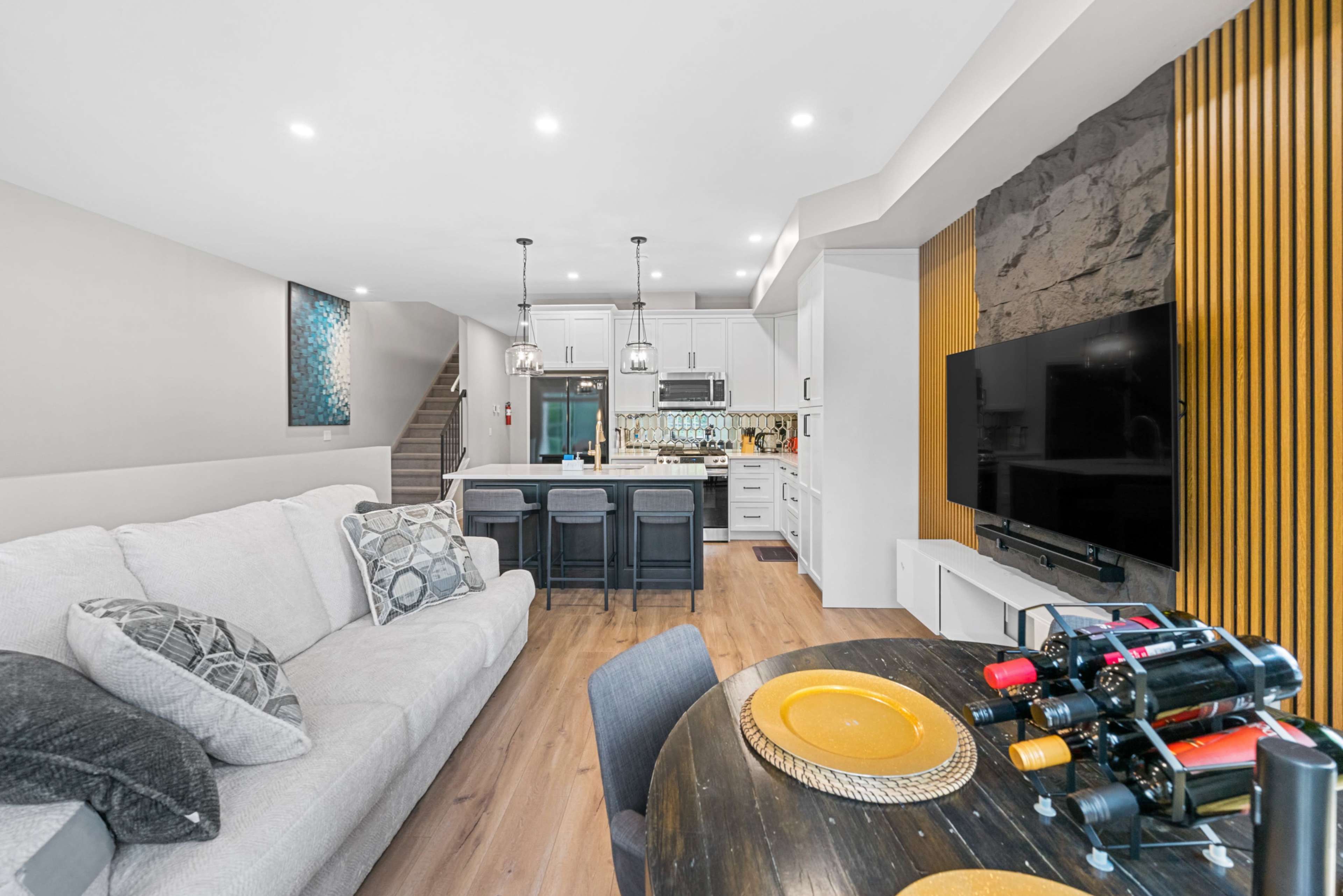 The image shows a modern living space featuring a sofa, a dining table with wine bottles, and a kitchen visible in the background.