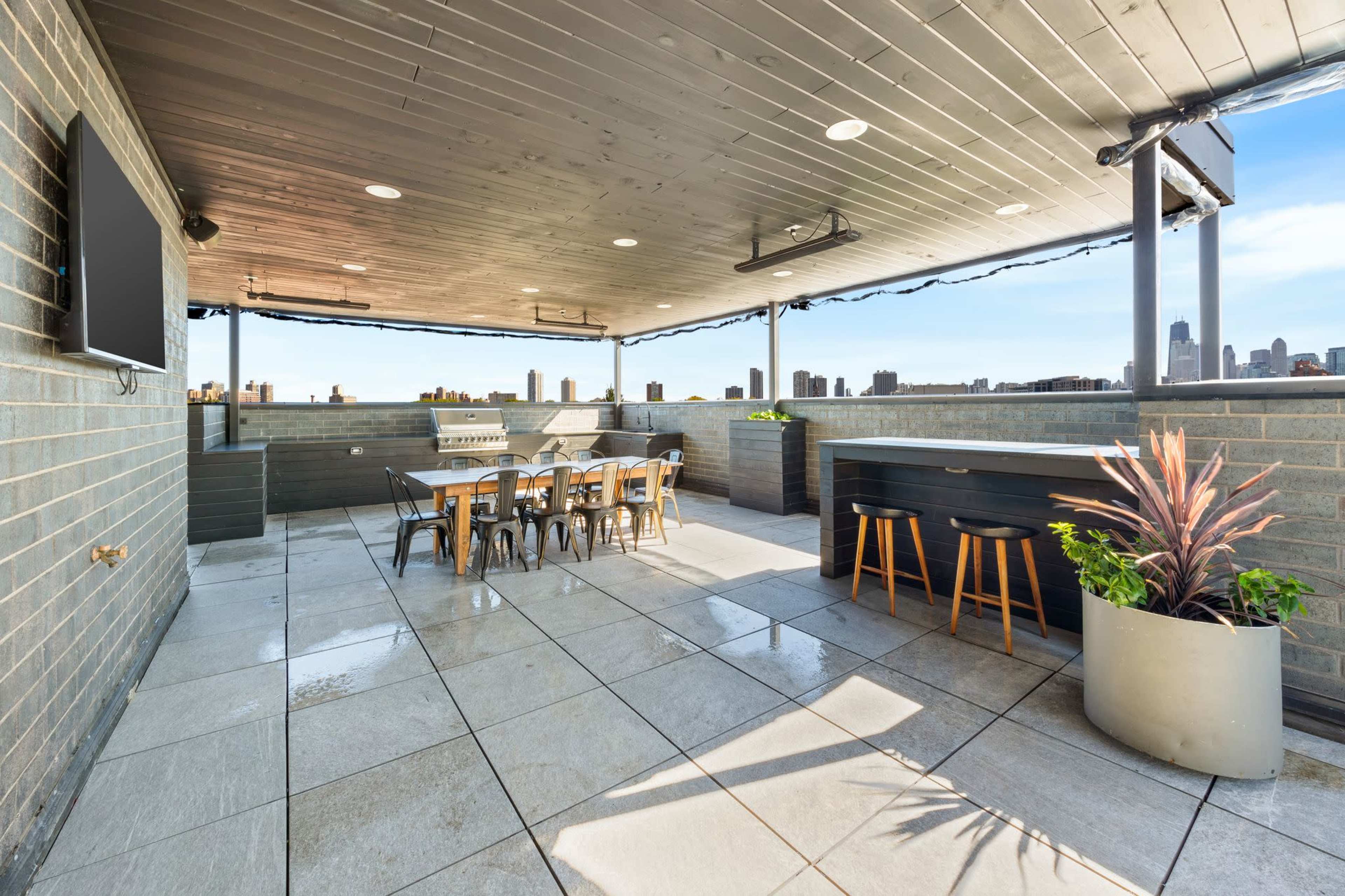 The image shows a spacious rooftop terrace with a dining area, outdoor bar, and skyline views of a city in the background.