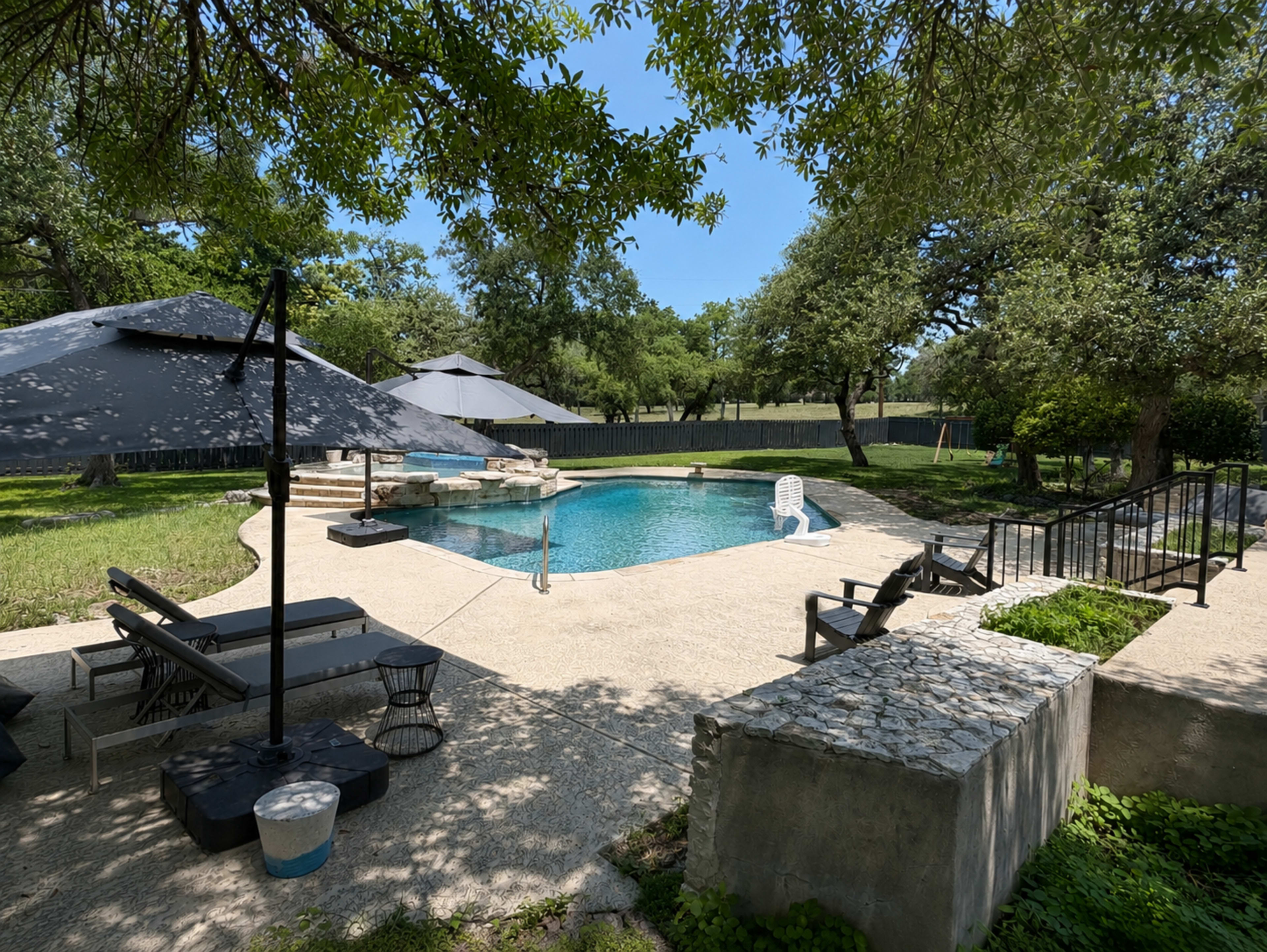 The image shows a backyard with a swimming pool surrounded by lounge chairs and shade umbrellas, set among trees and grassy areas.