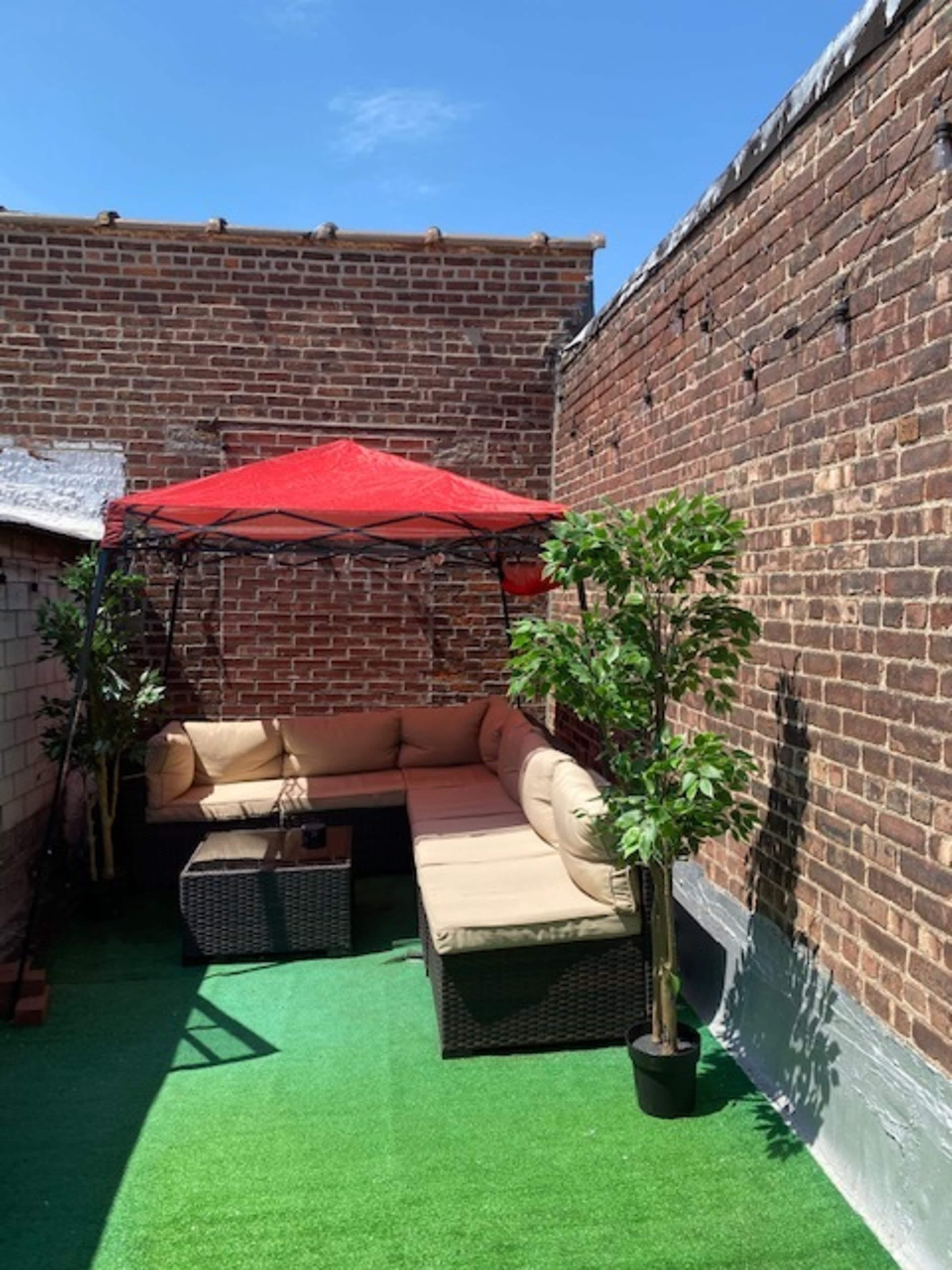 A rooftop terrace features a red canopy over a beige sectional sofa, surrounded by potted plants and artificial grass.