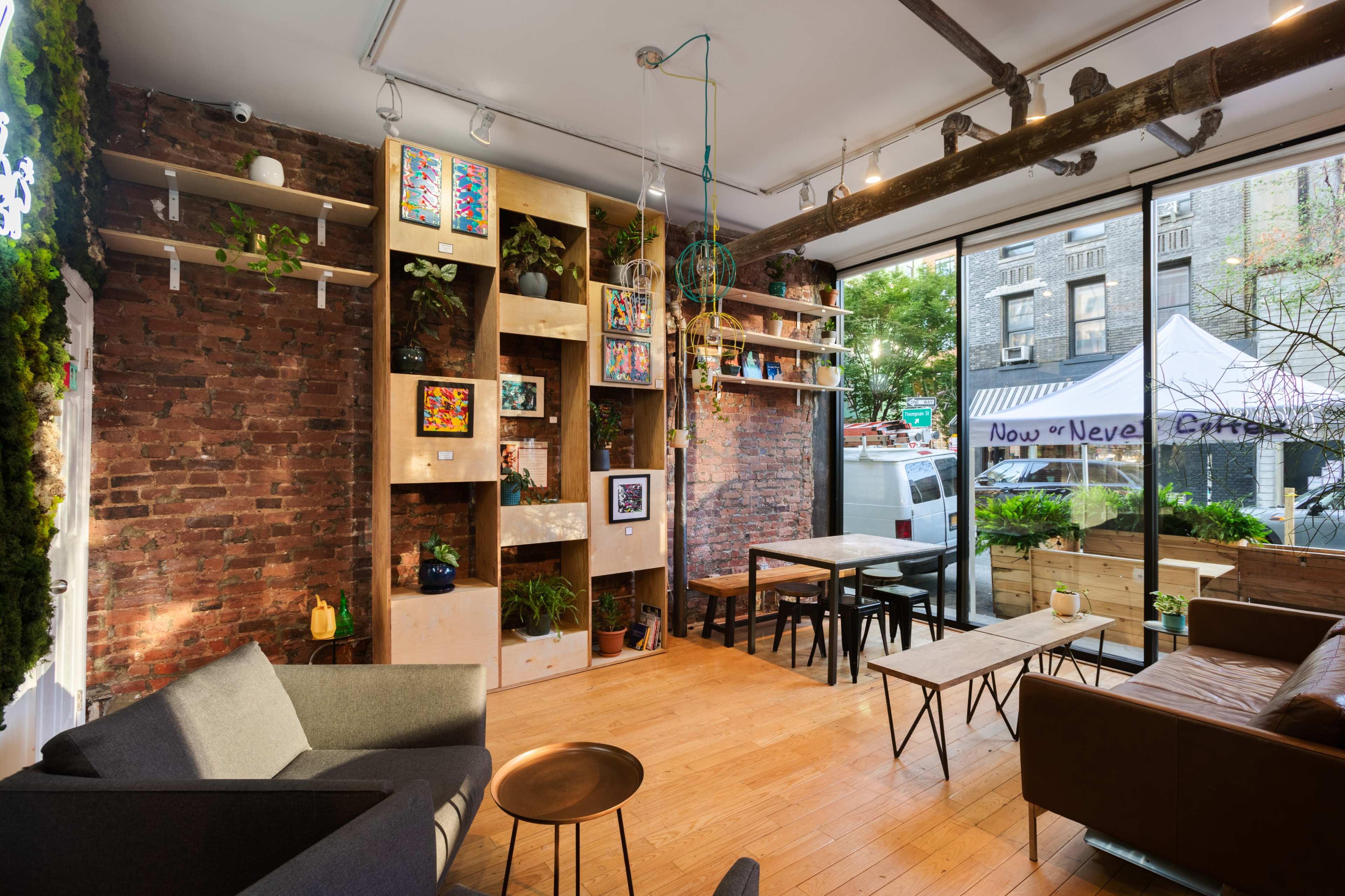 A cozy café features exposed brick walls, wooden shelving with plants and art, and multiple seating arrangements by large windows.