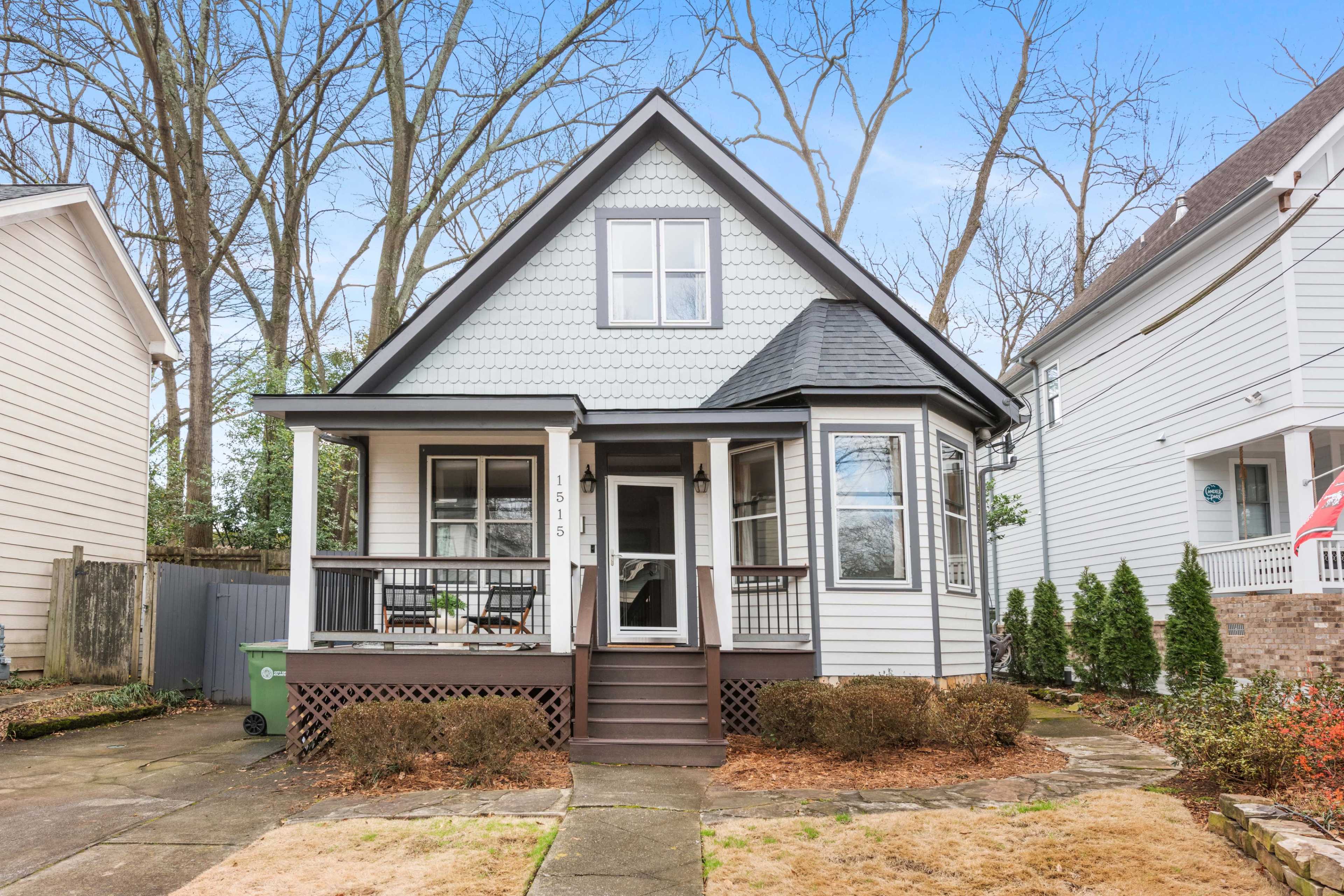 A charming two-story house with a steep roof and a front porch is set among bare trees in a suburban neighborhood.