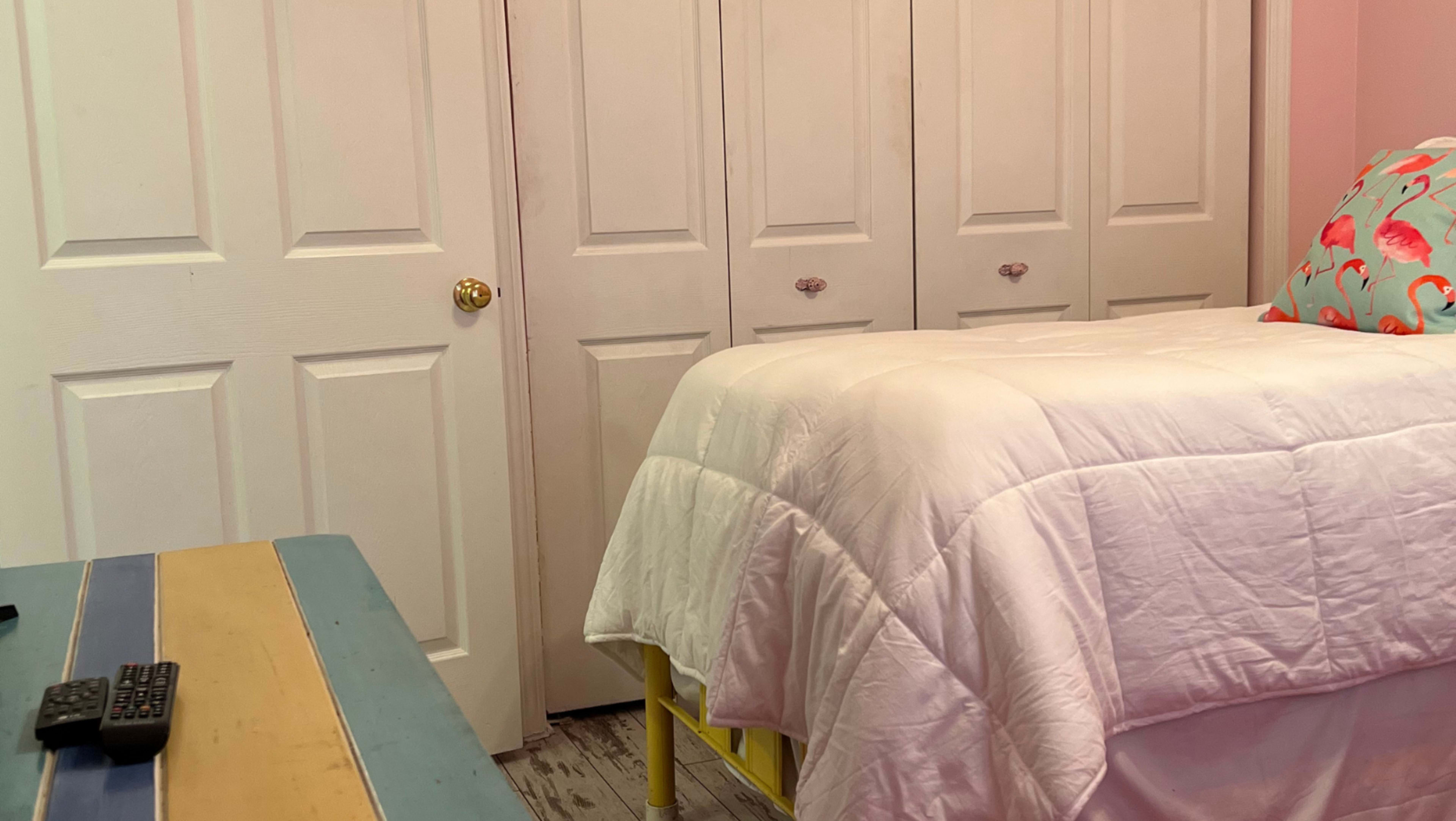 A neatly made bed with a white comforter is positioned beside a blue and yellow striped table in a room featuring light pink walls and closed closet doors.