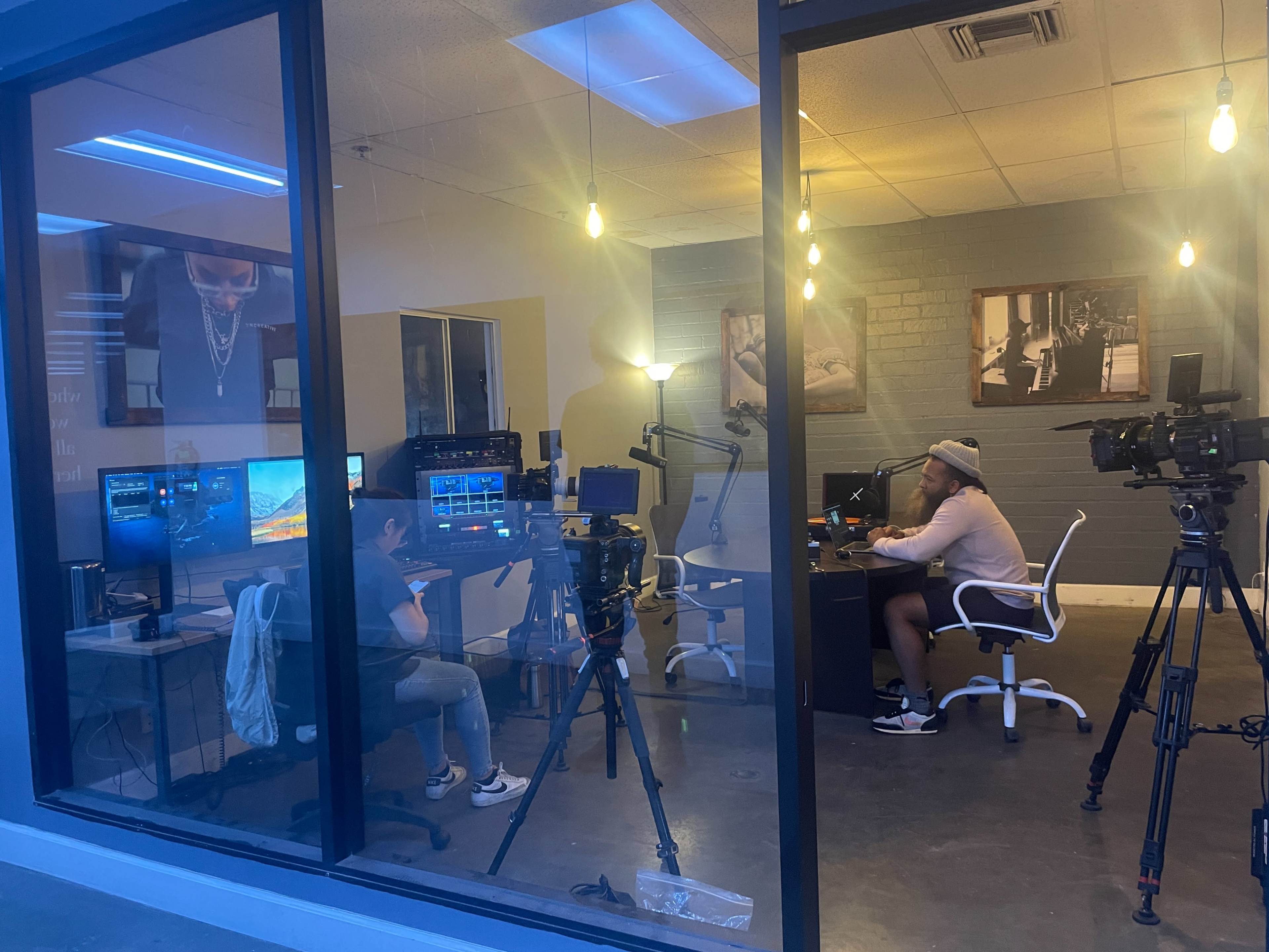 A media studio features two individuals set up for recording, with multiple cameras and monitors present in a well-lit environment.