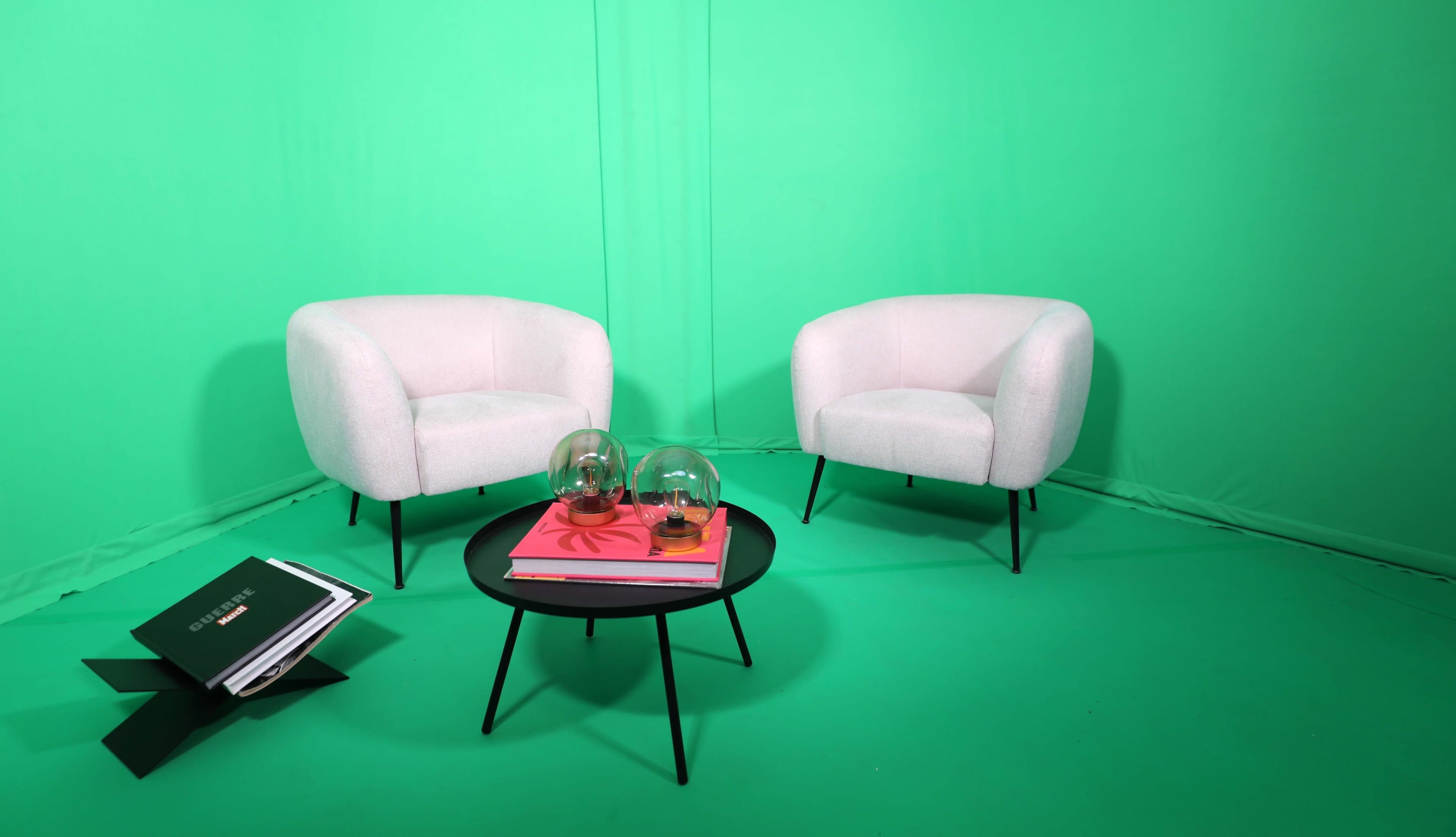 Two white armchairs face a black coffee table on a green backdrop, with a stack of magazines and glass globes arranged on the table.