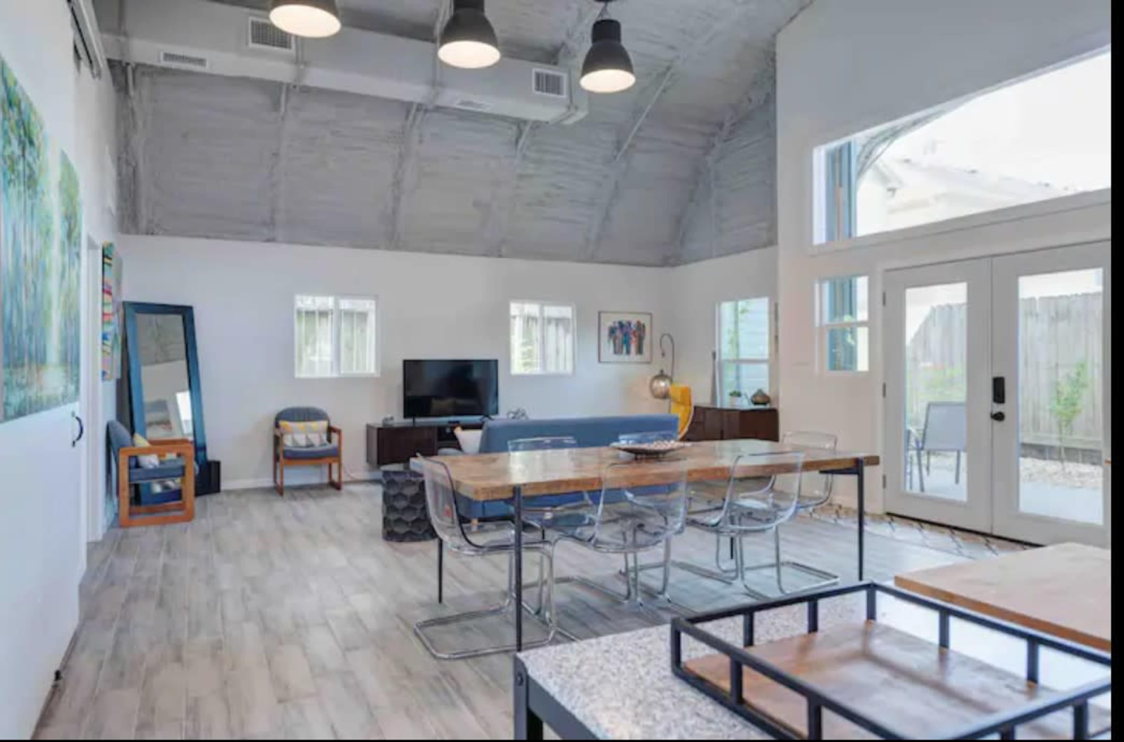 A spacious, modern living area featuring a large dining table, a television, and large windows that provide natural light.